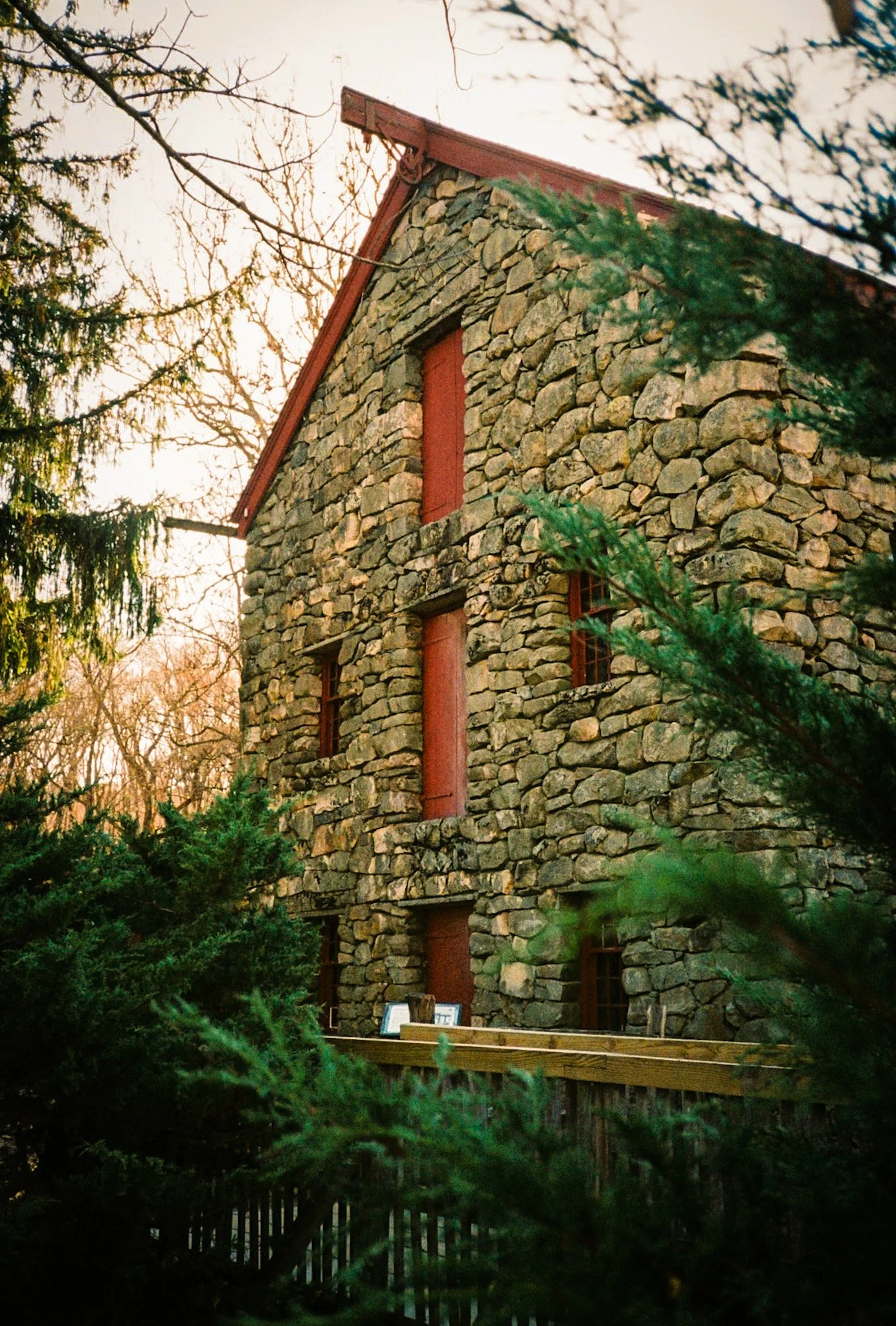 A stone house with small rectangular red shutters on the windows, surrounded by green trees and shrubs, during sunset.