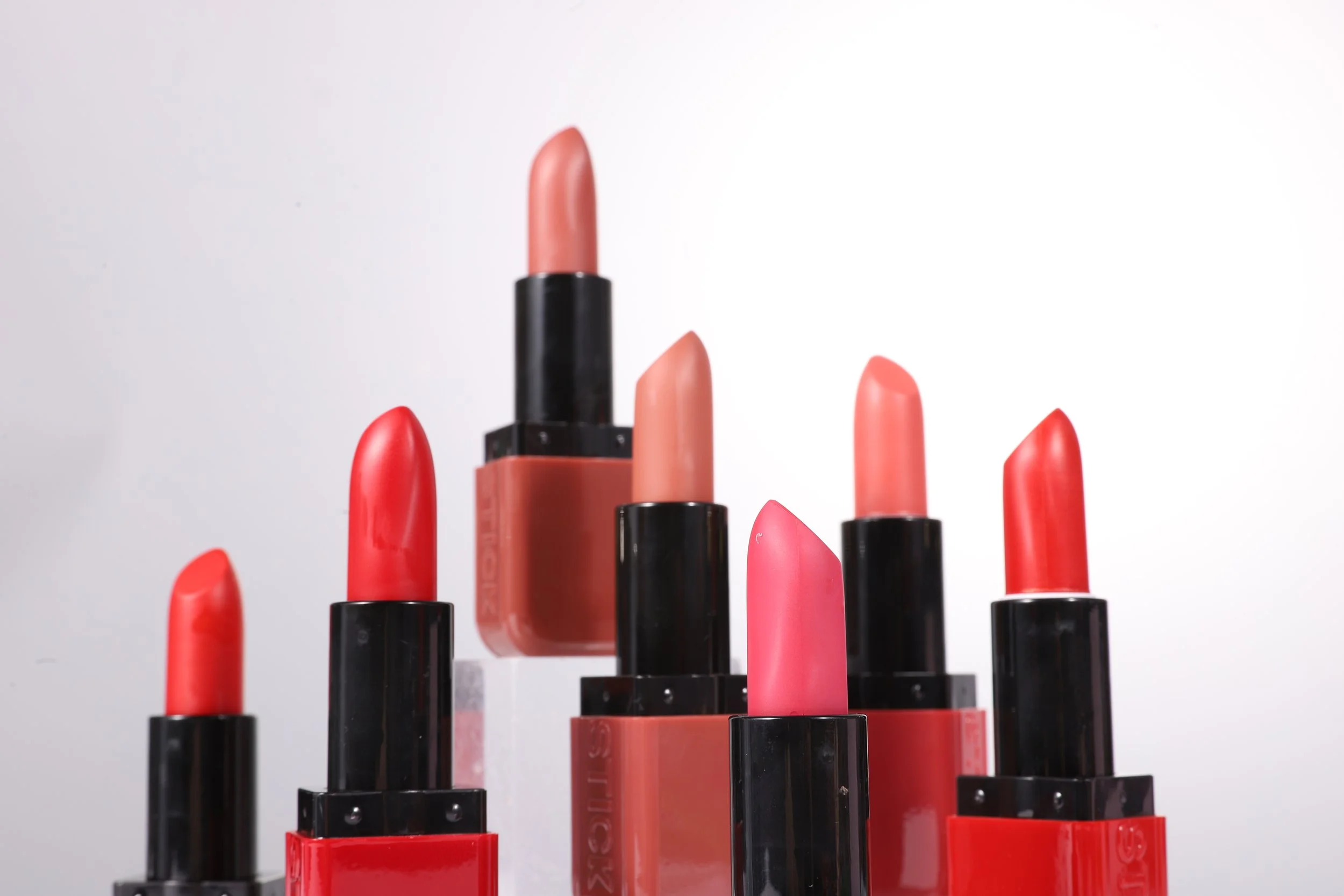 Vários batons em cores diferentes, dispostos em altura e direção variadas, contra fundo branco.