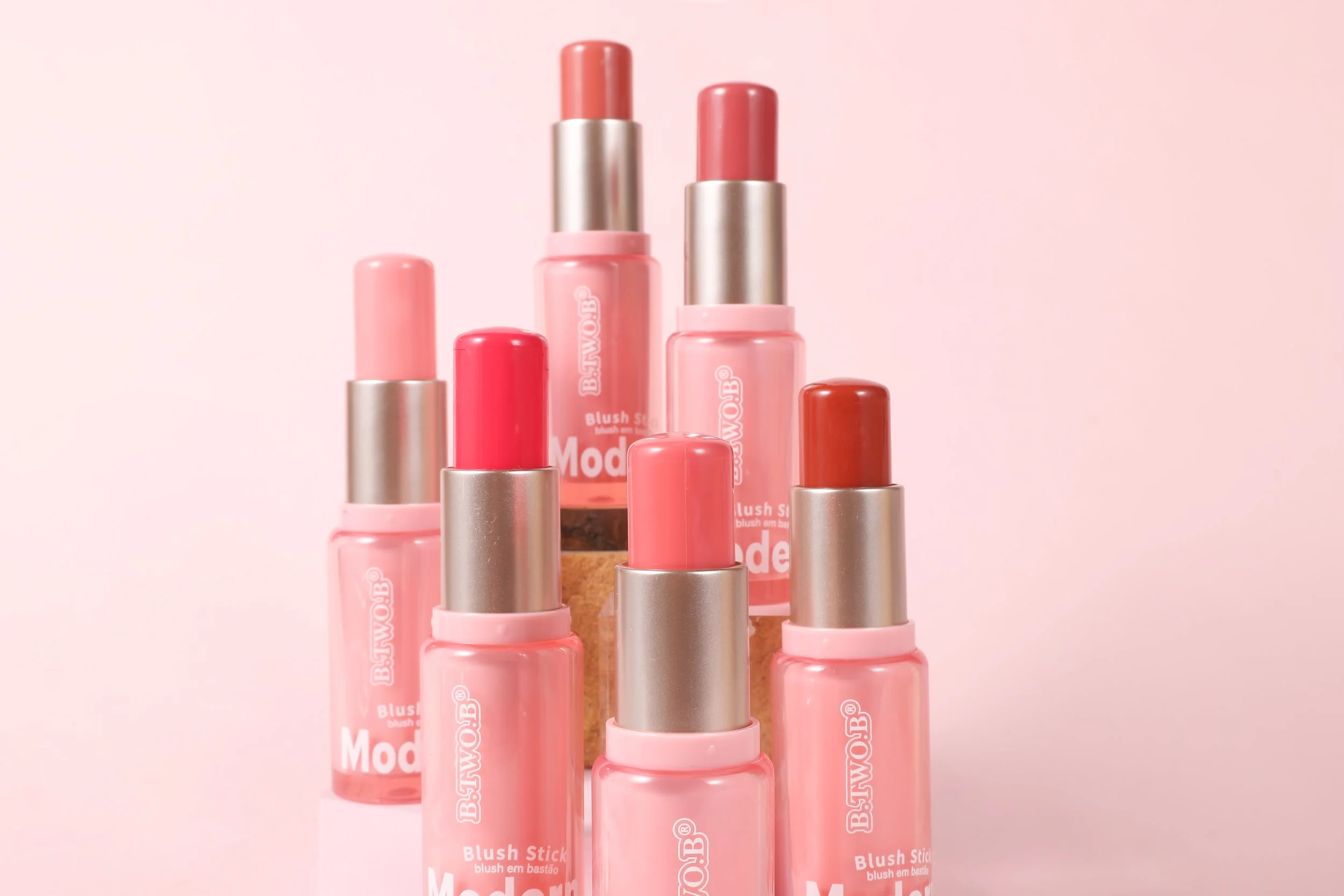 Vários batons em tons de rosa, vermelho e nude dispostos em uma escada com fundo rosa claro.