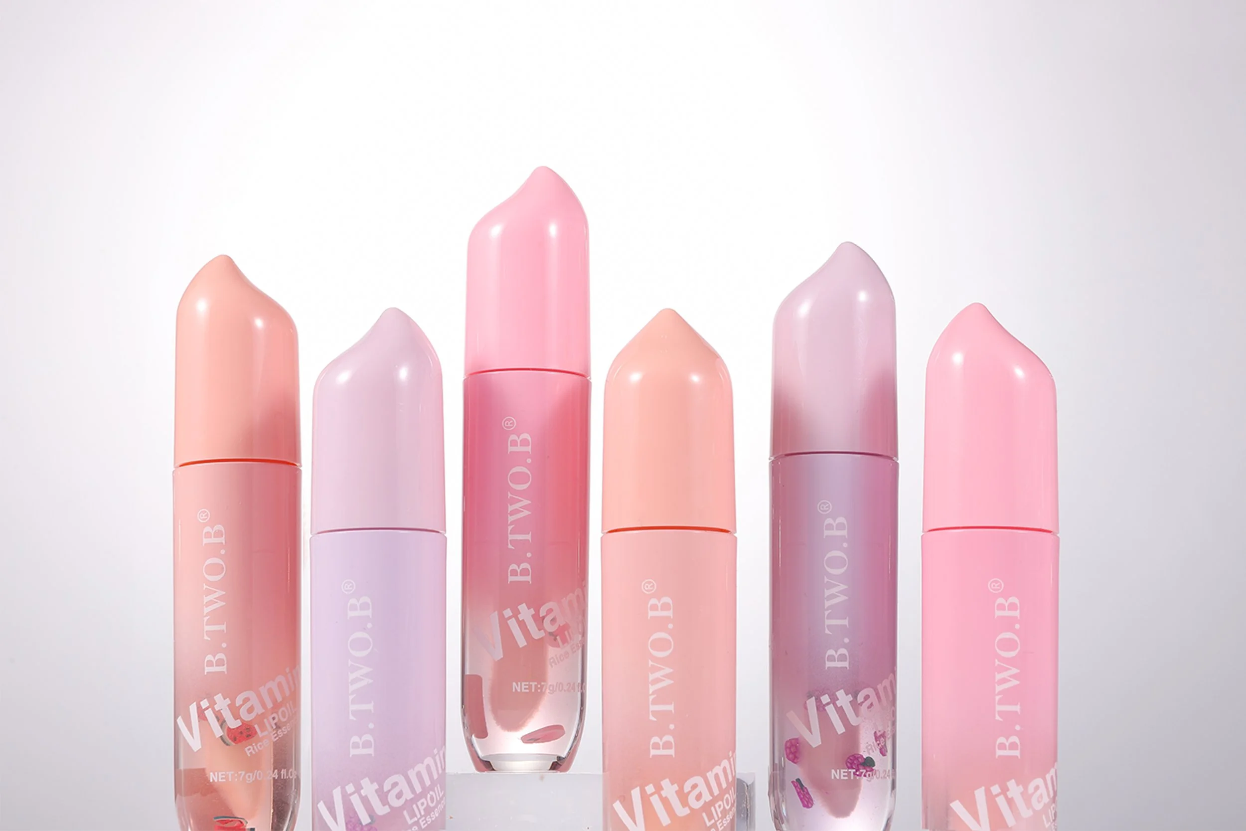 Seis batons de lip gloss de tons pastel variando de rosa claro a lilás, com embalagens translúcidas e com a palavra 'Vitamin' escrita na frente, posicionados em uma superfície branca com fundo cinza.