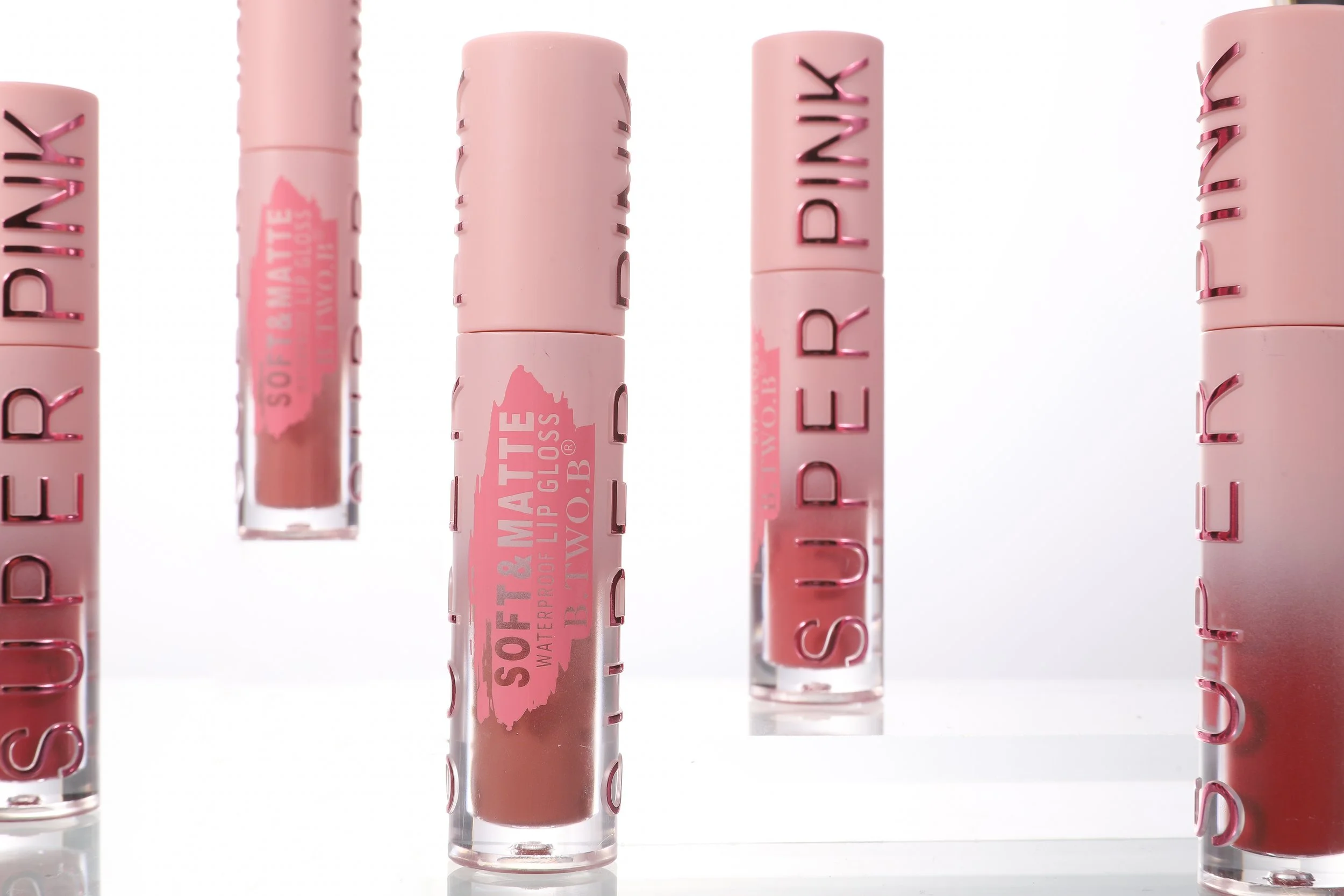 Variedade de batons líquidos em diferentes tons de rosa e nude, com embalagem transparente e detalhes em rosa escuro, exibidos contra fundo claro.