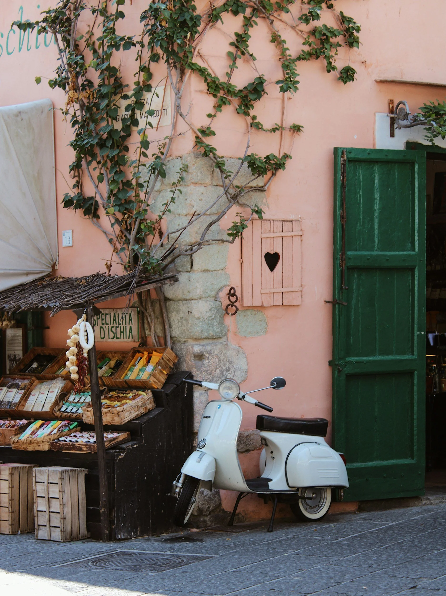 White scooter parked next to a small outdoor shop with colorful packaged goods, against a pink wall with a small green window shutter and green plants climbing the wall.