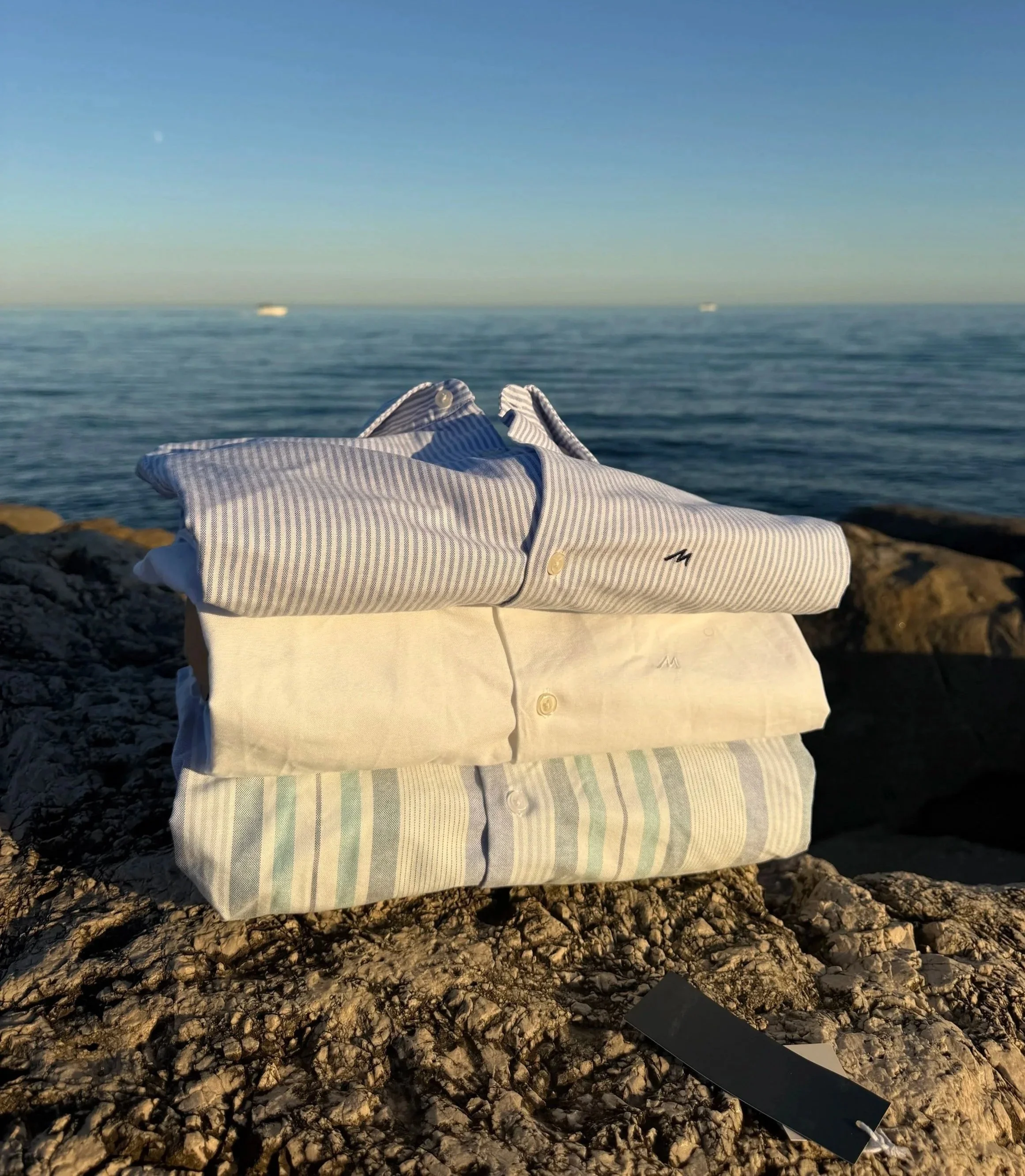 Three neatly folded striped cutaway shirts placed on rocky terrain by the Mediterranean sea with boats on the water in the background.