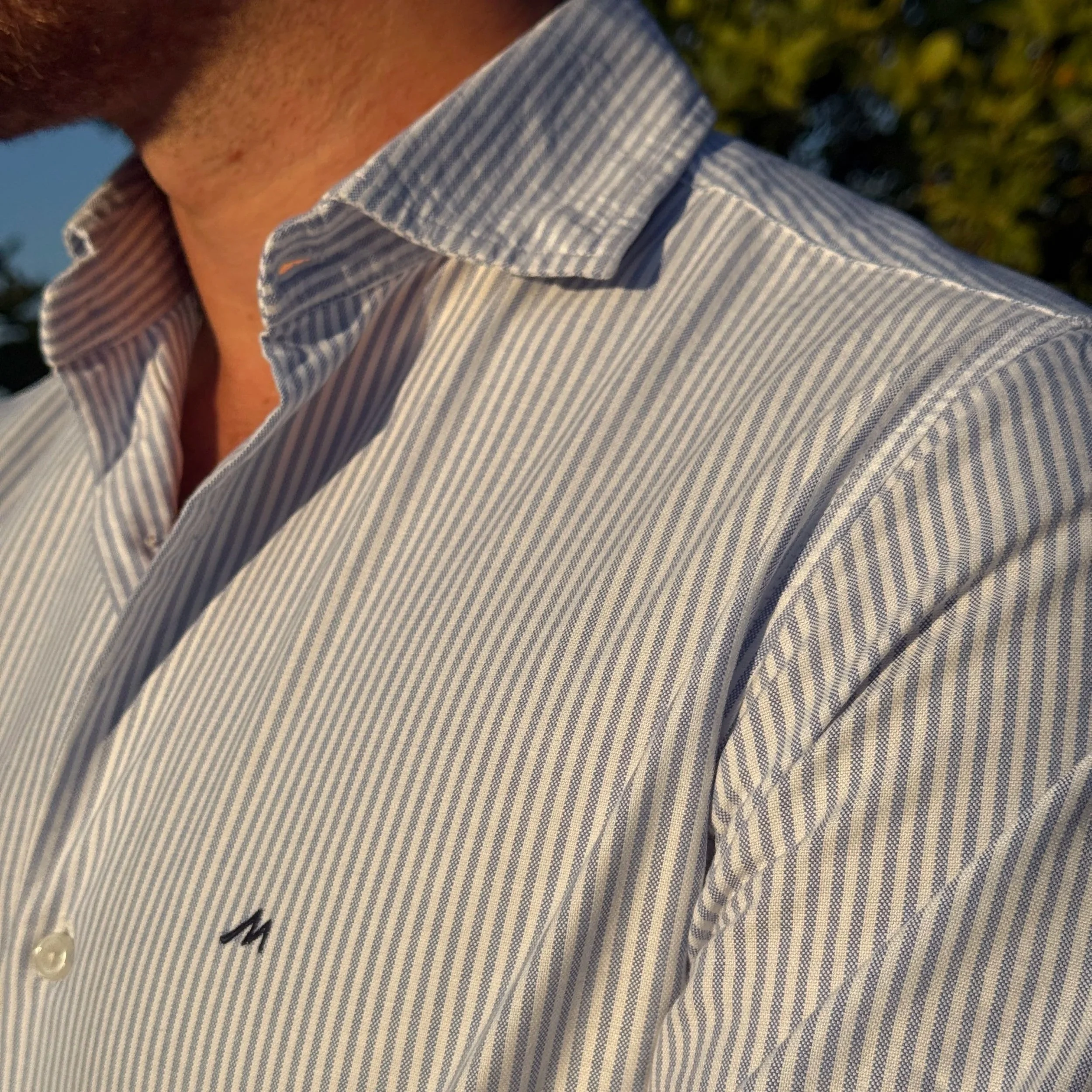 Close-up of a man wearing a light blue and white striped cutaway collar shirt, with the collar slightly open with the buttons undone, outdoors during daylight.