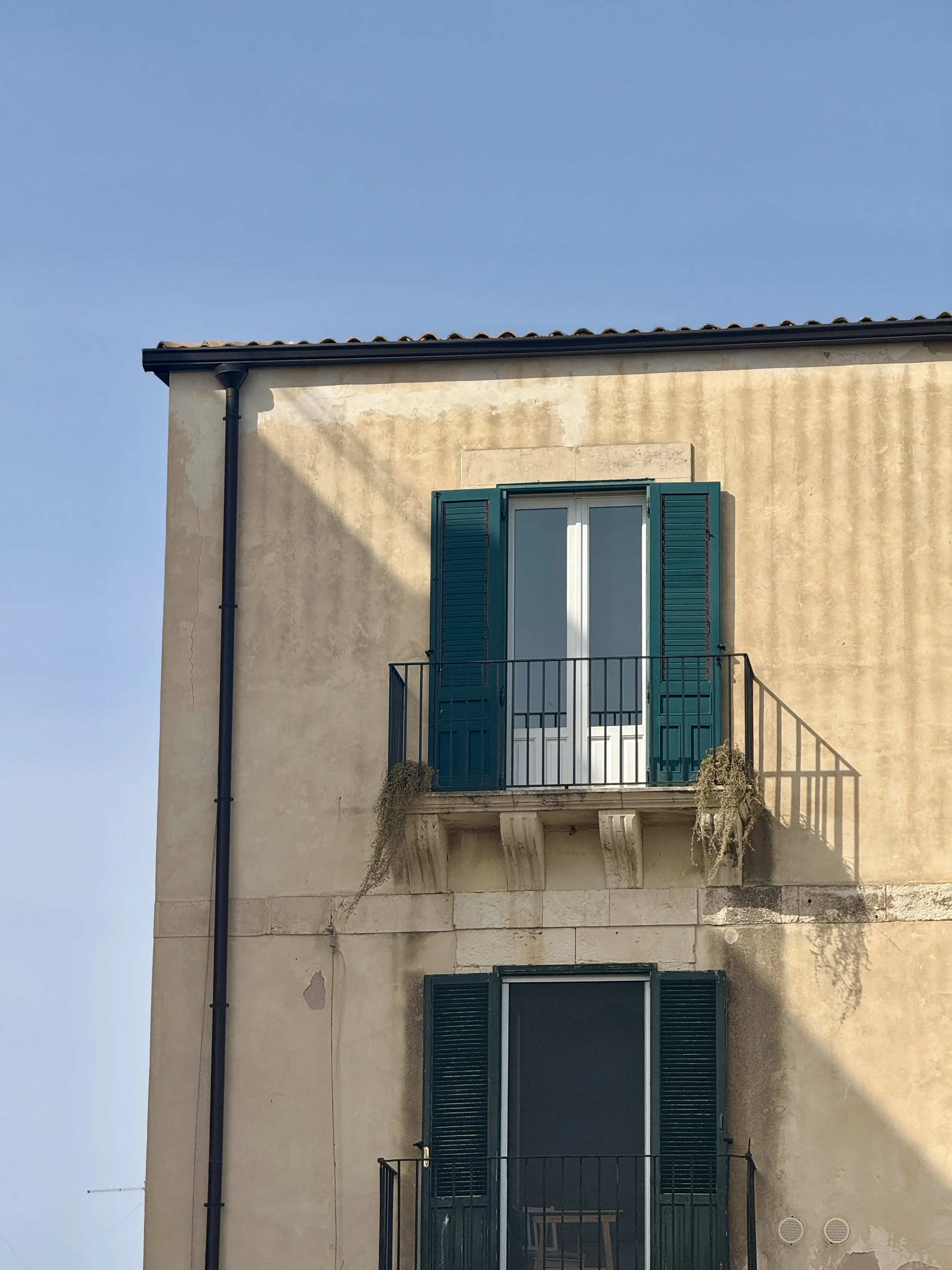 Photo of a building with two rustic italian balconies, one on top of the other, with teal shutters, black railings, and beige walls. The upper window is closed, with some plants hanging from the balcony.