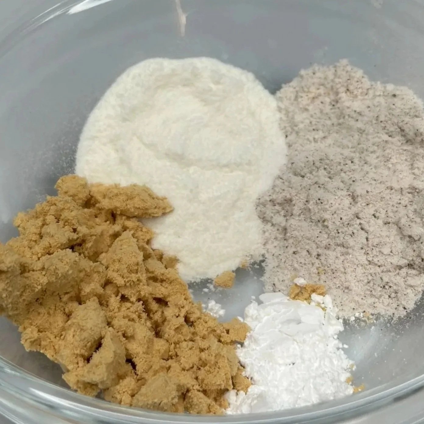 Assorted dry baking ingredients in a glass bowl, including flour, brown sugar, powdered sugar, and a light-colored ground spice or flour