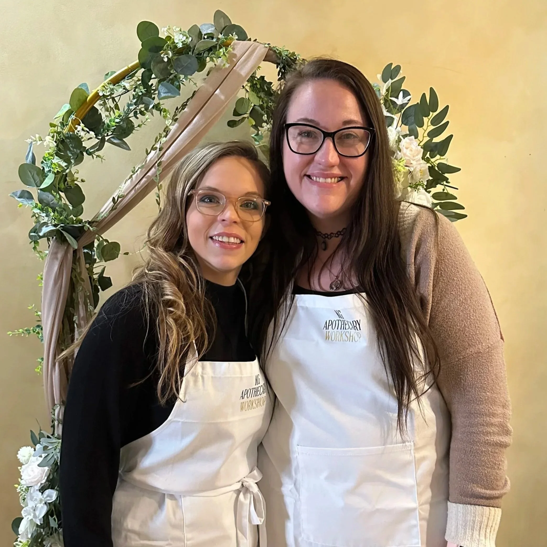 Two women smiling and standing close together, wearing aprons, in front of a decorative floral arch. One woman has long, wavy blonde hair and glasses; the other has long, straight dark brown hair and glasses. They are at an event called 'The Apothecary Workshop'.