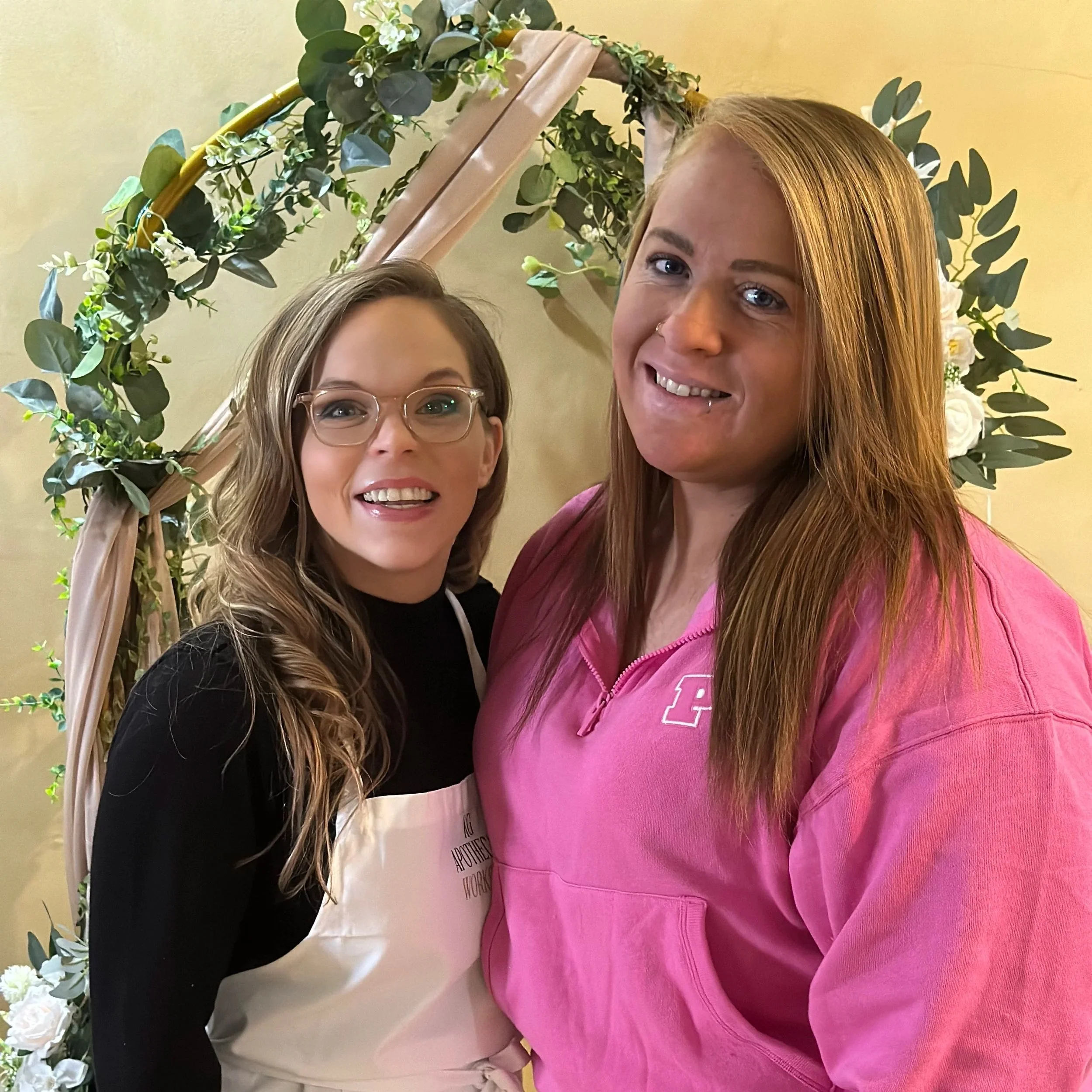 Two women smiling in front of a floral arch, one wearing glasses and a black top with an apron, and the other in a pink sweatshirt.