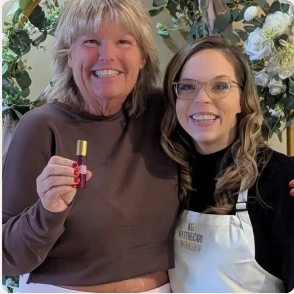 Two women smiling, one holding a small bottle of perfume or essential oil, with floral decorations in the background.