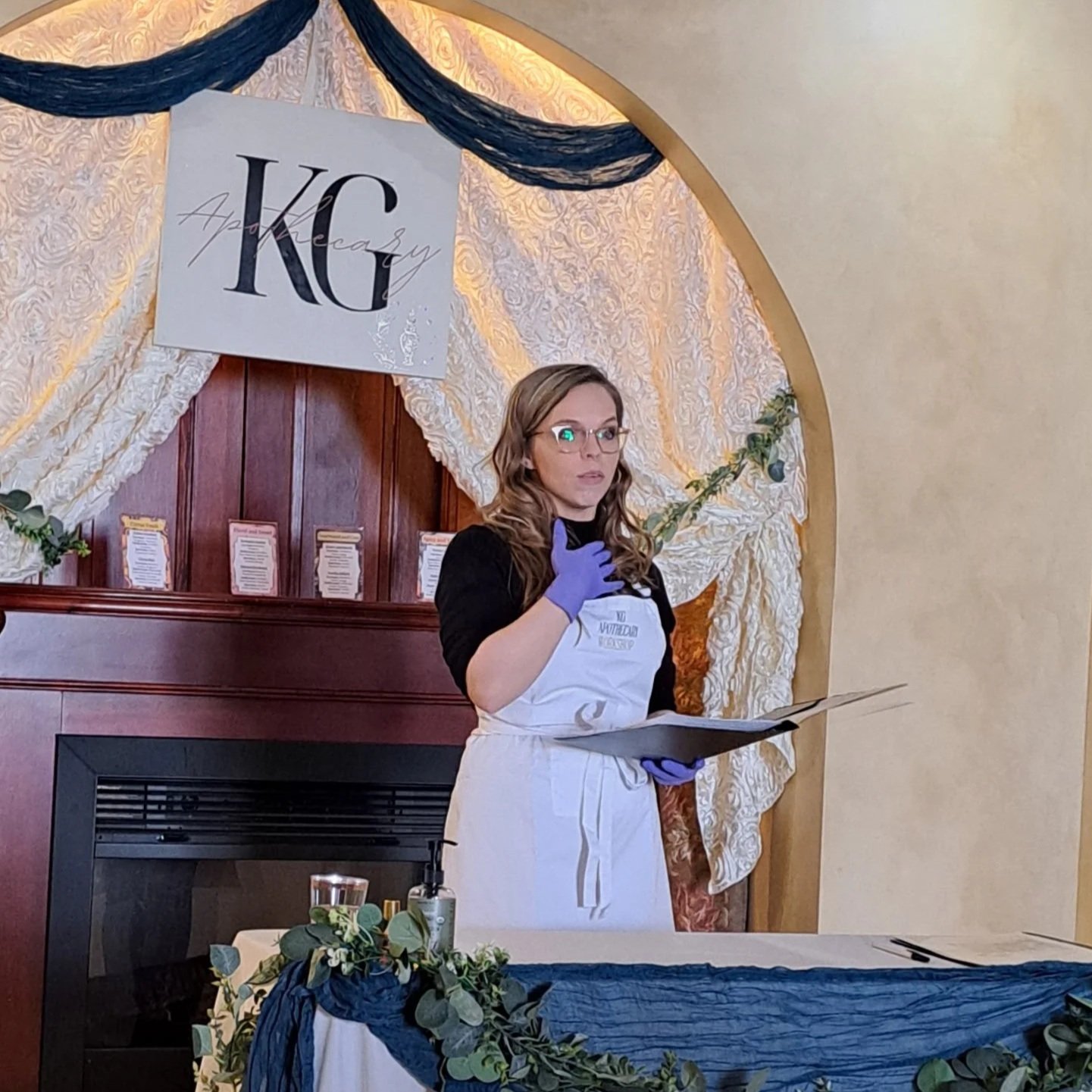 A woman wearing glasses, a black shirt, purple gloves, and a white apron stands in front of a decorated backdrop with a sign that reads 'KC Apacary'. She holds a clipboard and is gesturing with her right hand. The setting appears to be an event or presentation space with a table decorated with greenery and a candle in the foreground.