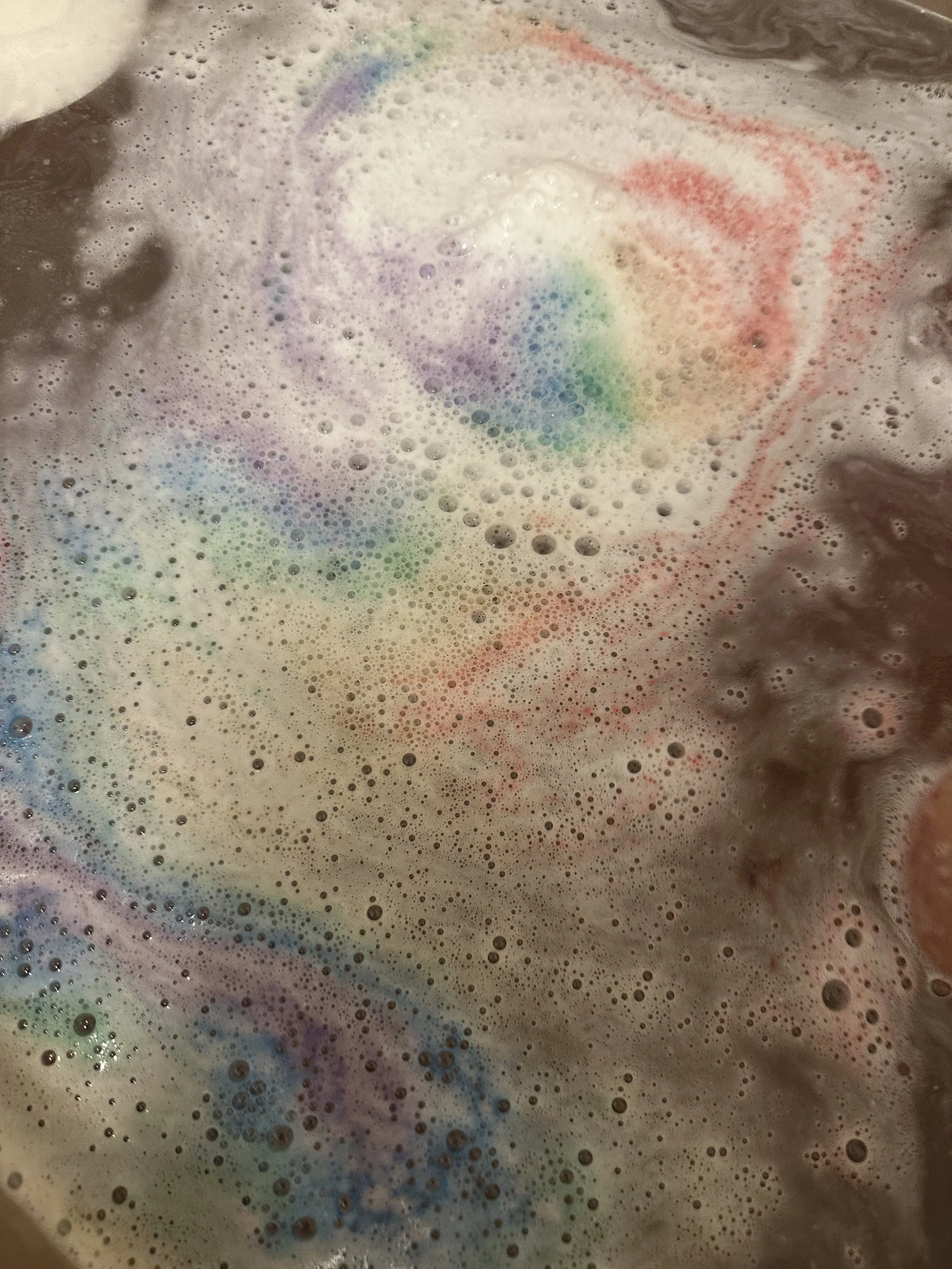 Colorful swirling bath bomb dissolving in water, creating foam and bubbles with rainbow hues.