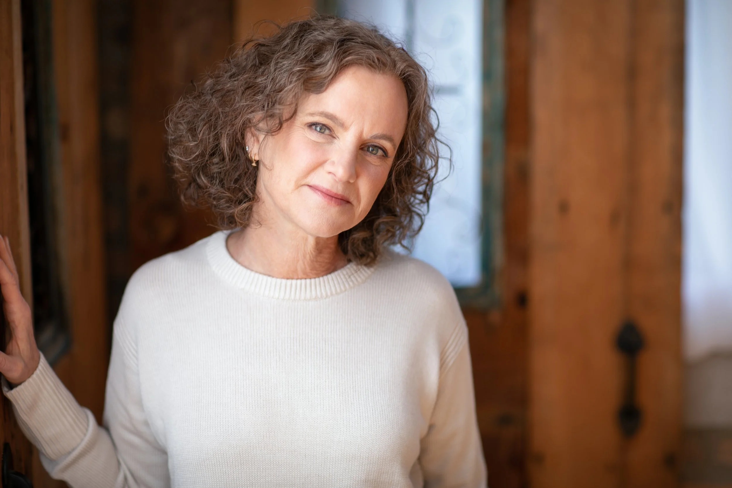 A woman with curly brown hair and blue eyes wearing a white sweater, standing in front of wooden walls, with a slight smile on her face.