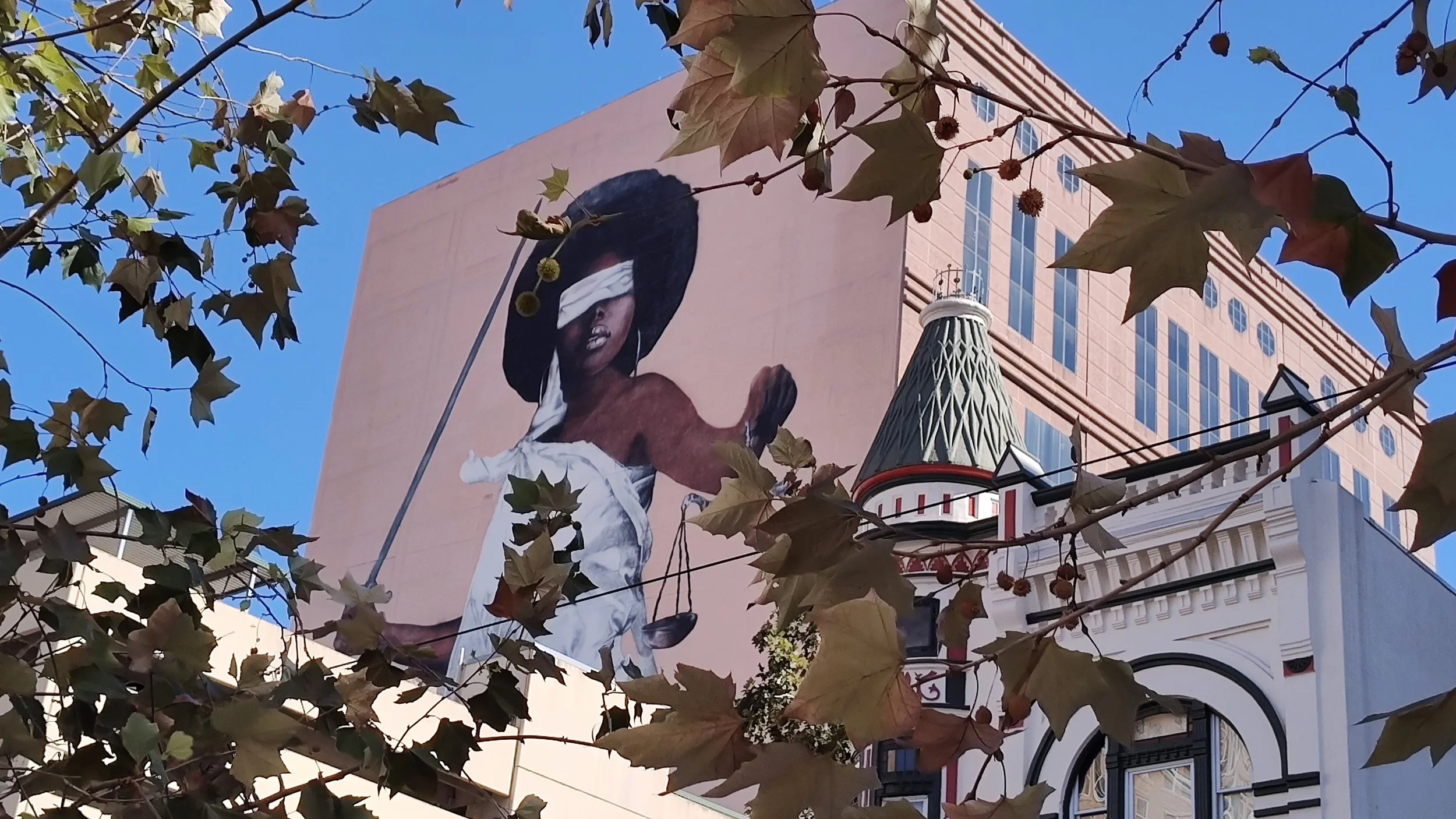 Large mural of a person with dark hair covered with a blindfold, shirtless, holding a flagpole, on the side of a building, with tree branches and leaves in the foreground, and a clear blue sky in the background.