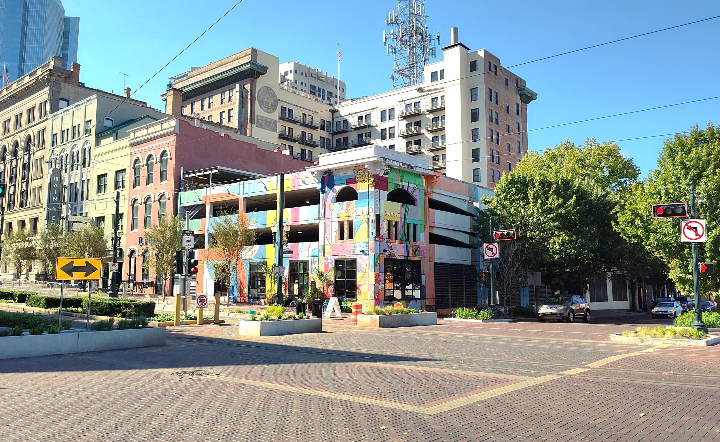 City street scene with colorful mural on building, trees, cars, traffic signals, and multi-story buildings.