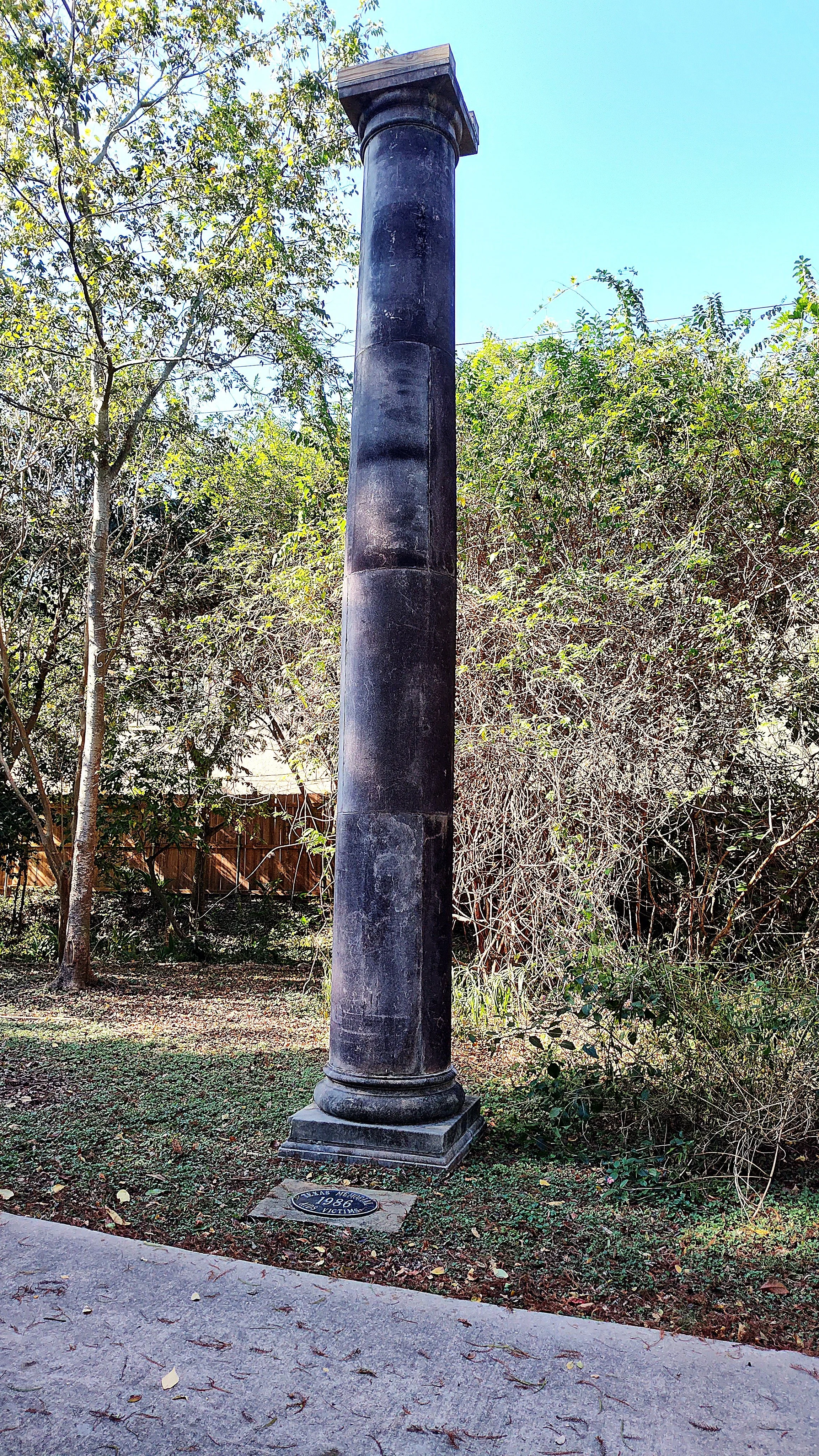 Historical black stone column with a plaque at its base, set in a garden with trees and bushes.