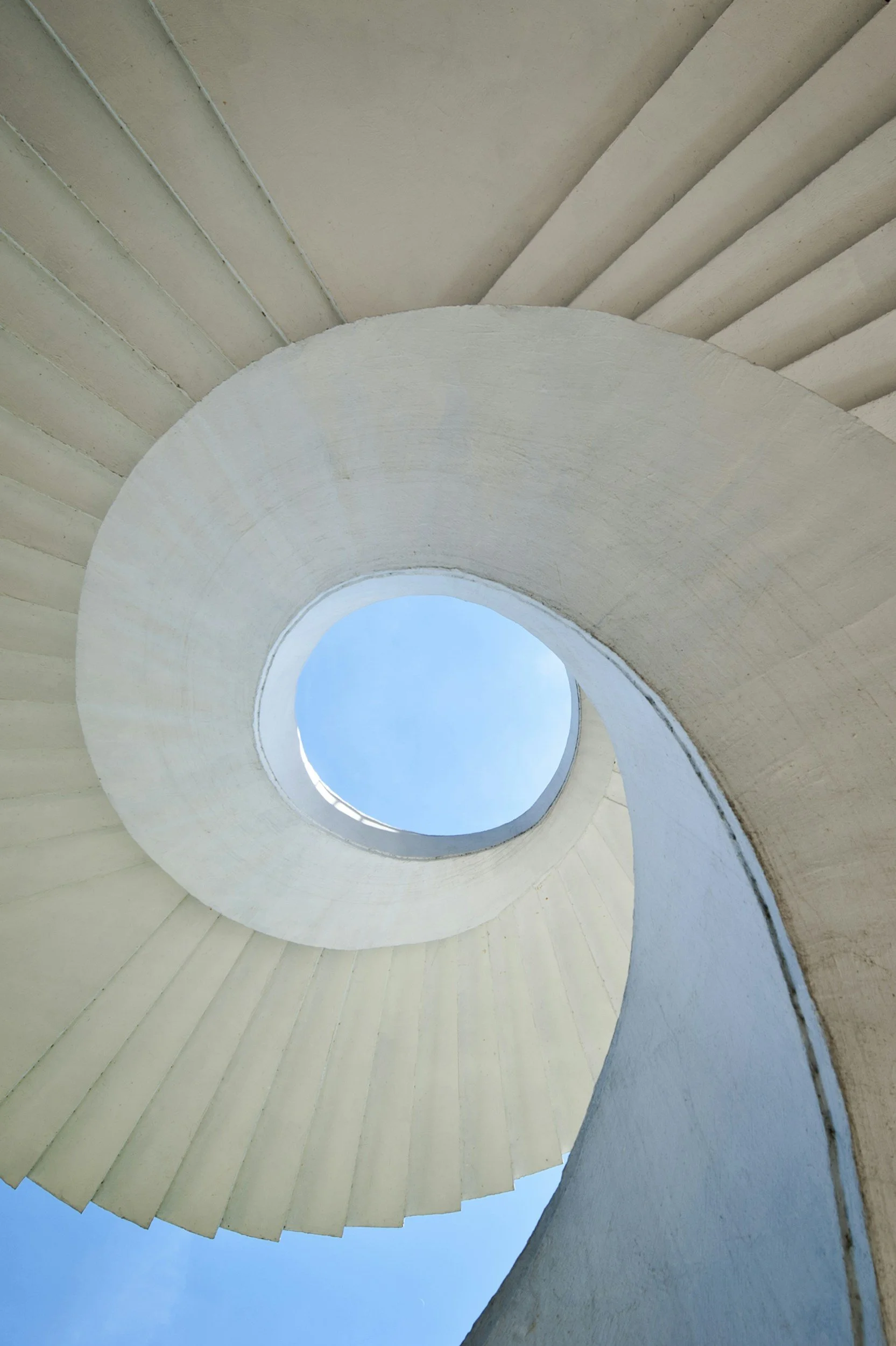 Vue du bas d'une escalier en colimaçon blanc vu du sol, avec le ciel bleu en arrière-plan.