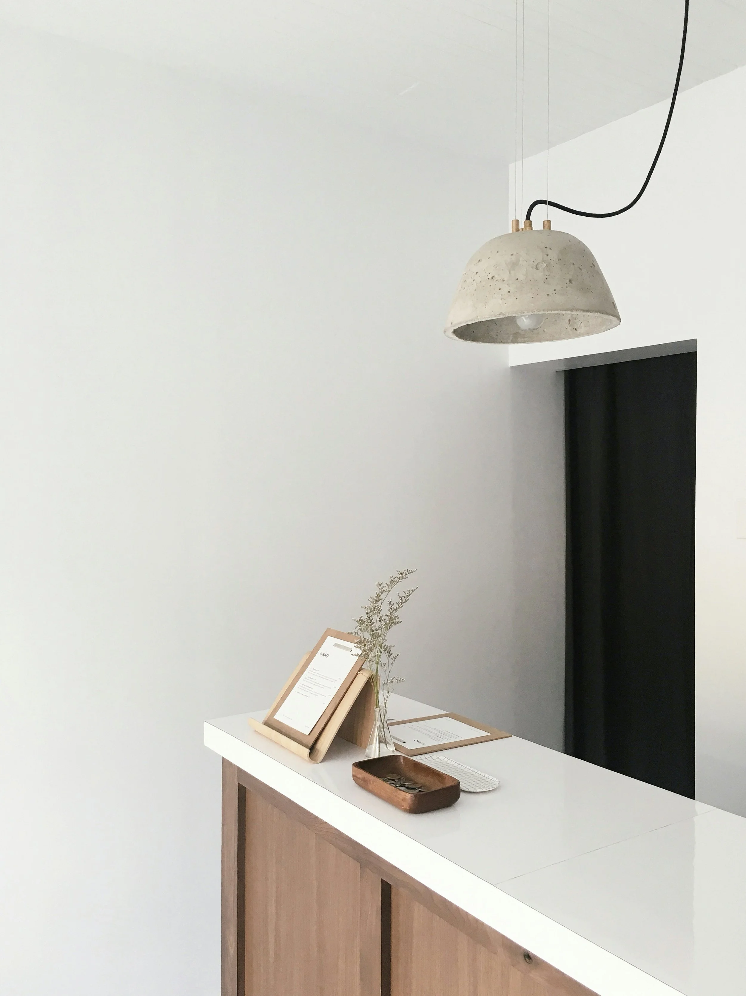 Minimalist reception desk with a white top, wooden base, a small vase with greenery, framed document, wooden tray, and a computer keyboard. A concrete pendant light hangs overhead in a white-walled room with a black door or window in the background.