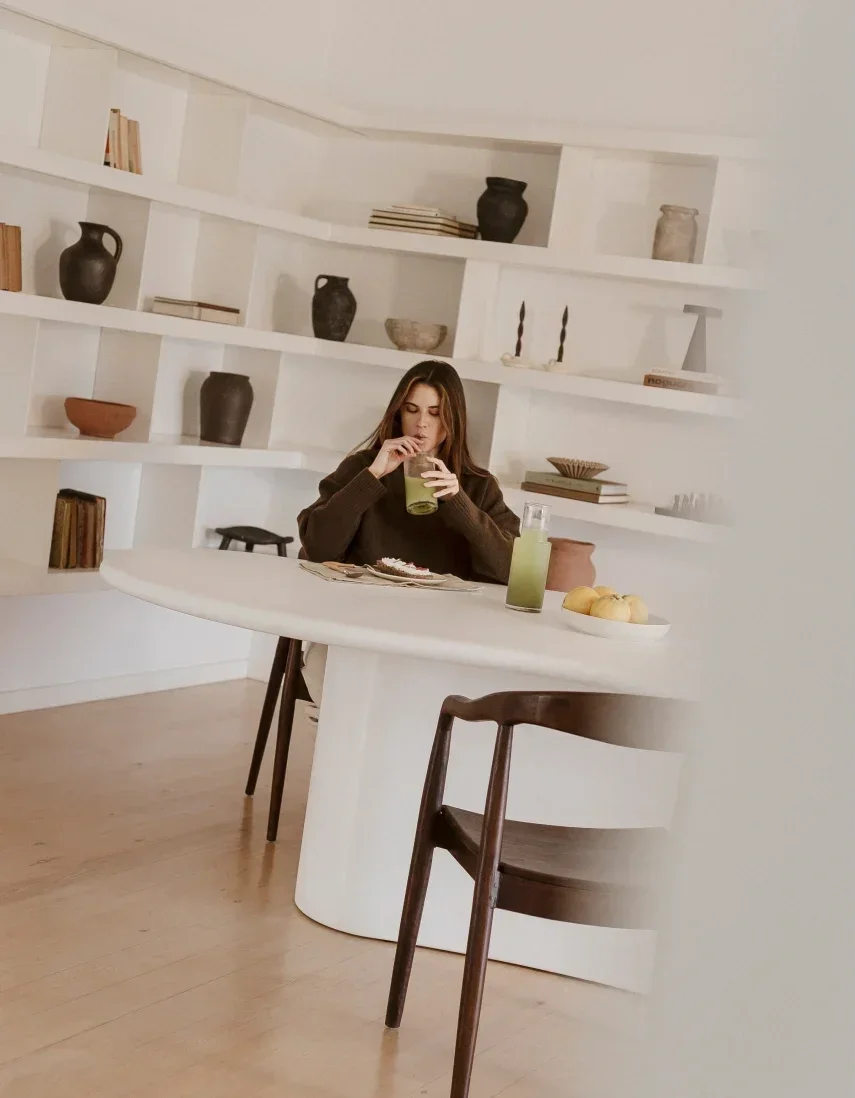 A woman sitting at a white dining table drinking a green beverage. There is a white plate with bread rolls and a tall glass with a matching green drink. The background features white shelves with decorative vases, books, and minimal decor.