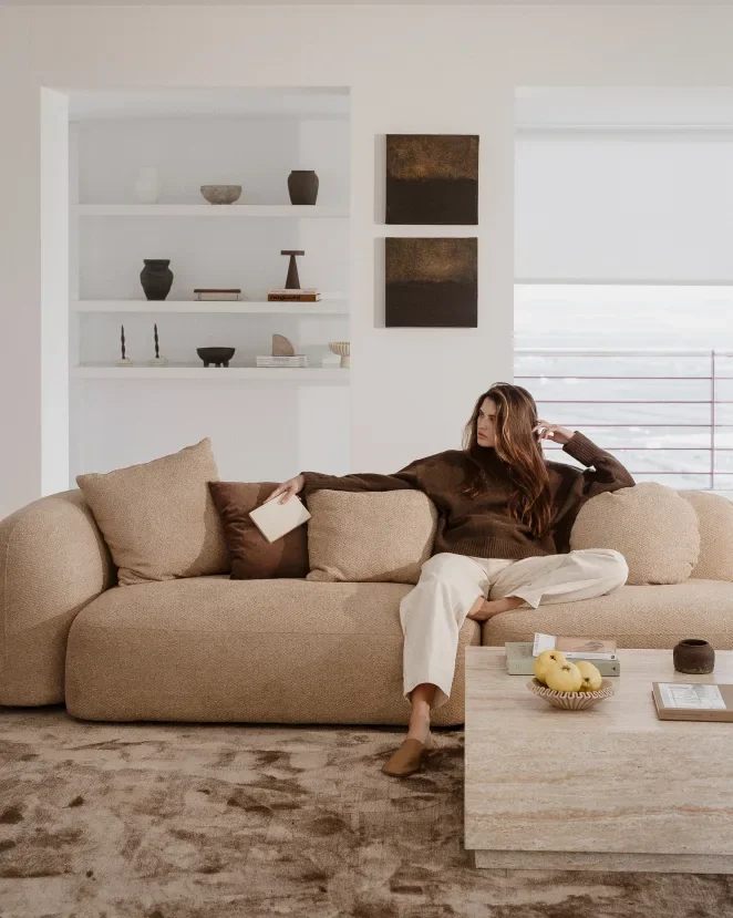 A woman sitting on a beige couch in a modern living room, holding a book with one hand, her legs crossed, dressed in a brown sweater and white pants. Behind her, white shelves with decorative objects and abstract artwork on the wall. A coffee table with a bowl of apples and a few books in front of the couch.