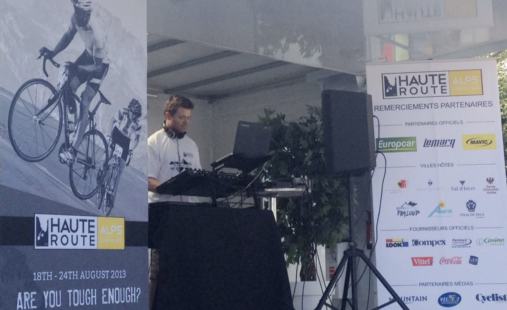 A male DJ playing music at a cycling event, with banners displaying event details and sponsors behind him.