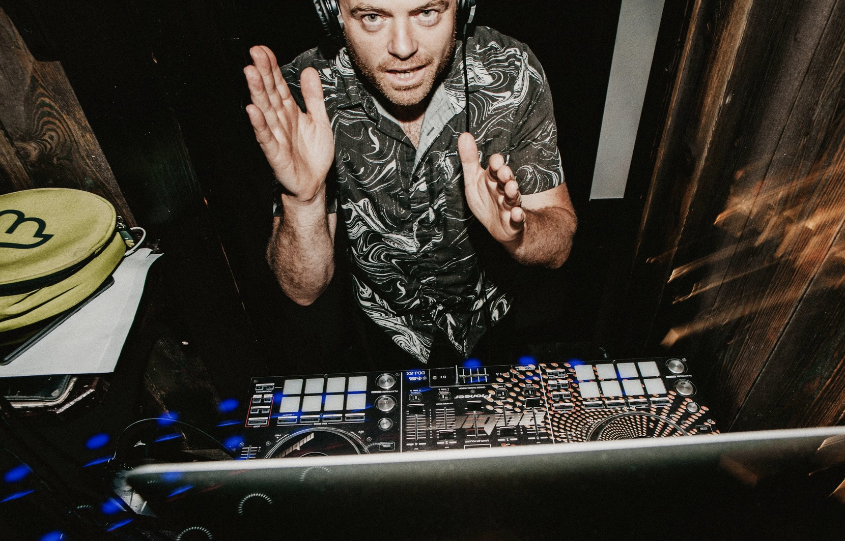 A male DJ wearing headphones and a patterned shirt is looking up while standing behind a DJ controller. He appears to be engaged with the music, with one hand raised near his face. The DJ setup includes a Pioneer DJ controller with various controls and illuminated buttons. There is a stack of yellow-green hats on the left side of the image, and the background consists of dark wooden panels.