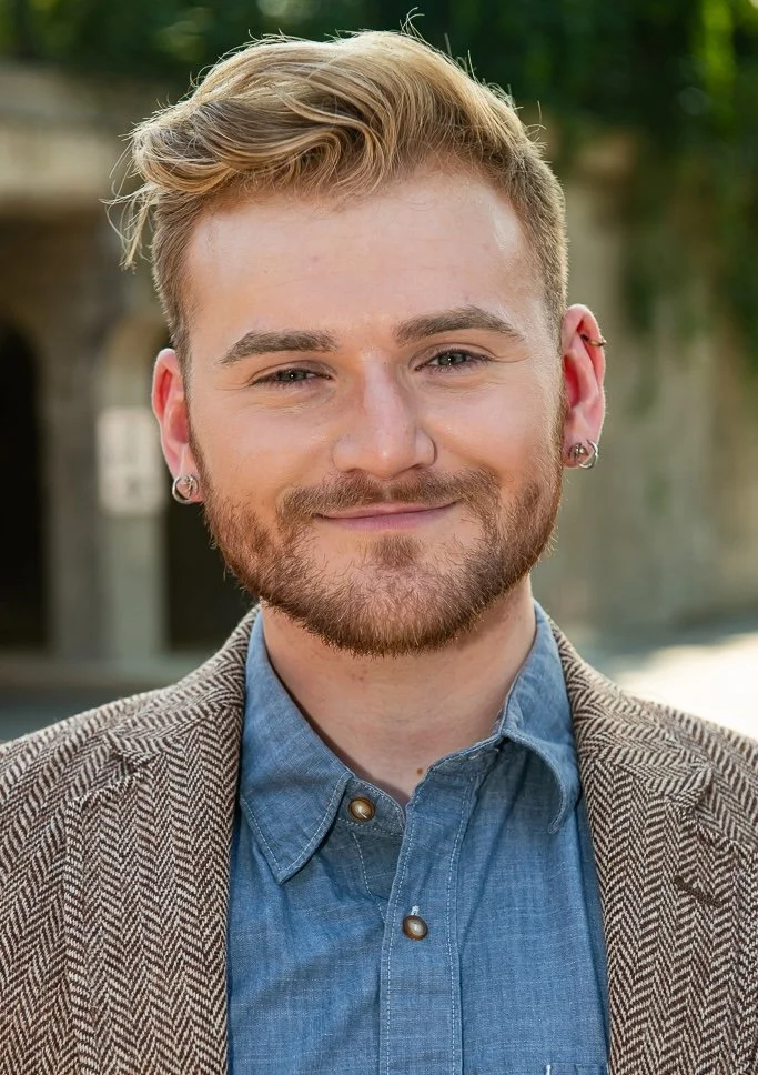 A young man with blonde hair and a beard, smiling, wearing a blue button-up shirt and a brown checkered blazer, outdoors with blurred trees in the background.
