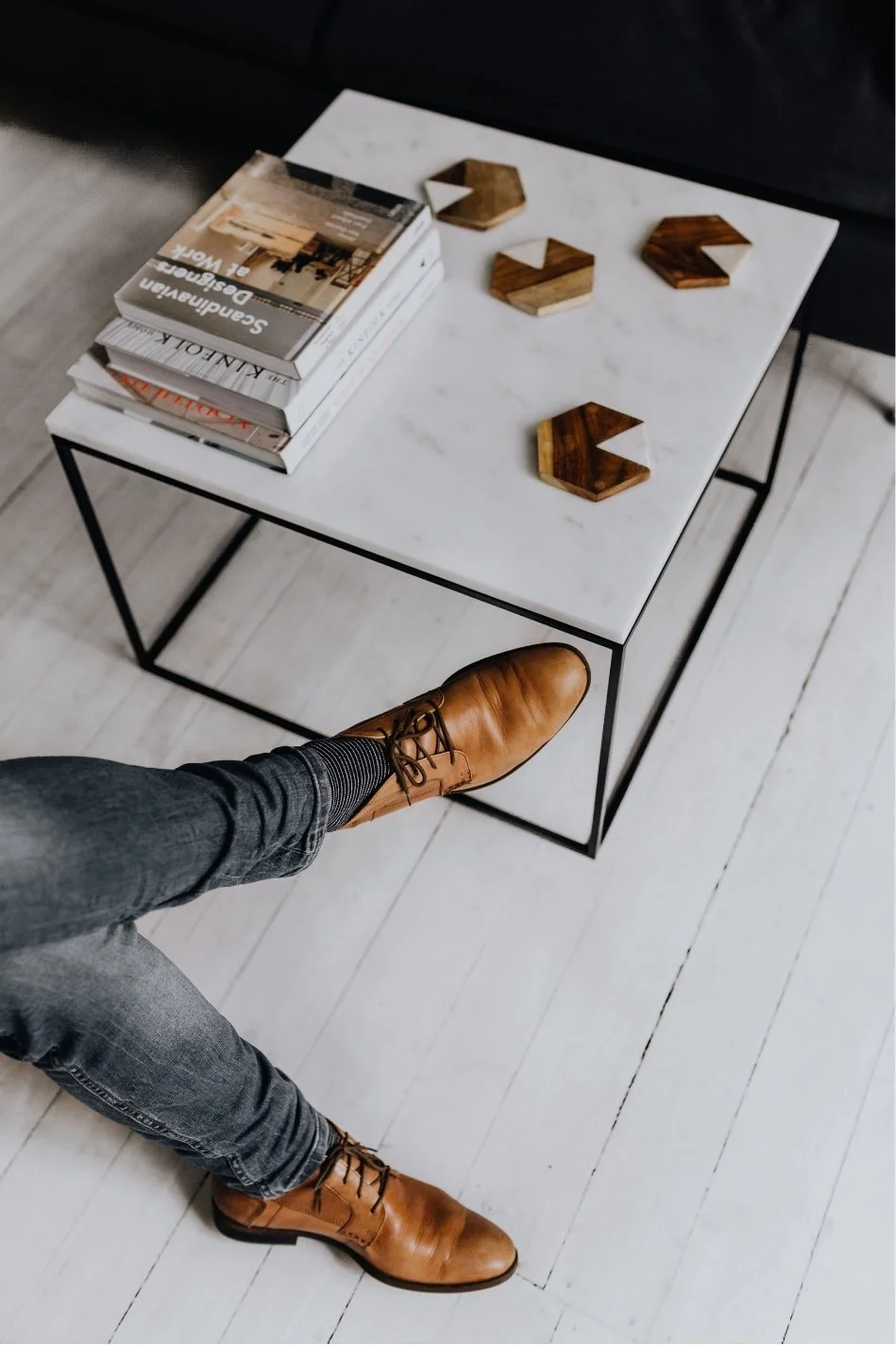 A white marble-top side table with black metal frame holds stacked books and wooden decorative coasters. A person wearing brown leather shoes, dark pants, and striped socks is sitting nearby on a white wooden floor.