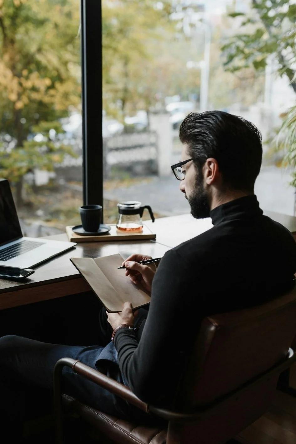 A man with glasses and dark hair, sitting by a window in a cafe, writing in a notebook with a pen, with a laptop, a coffee pot, and a cup on the table.