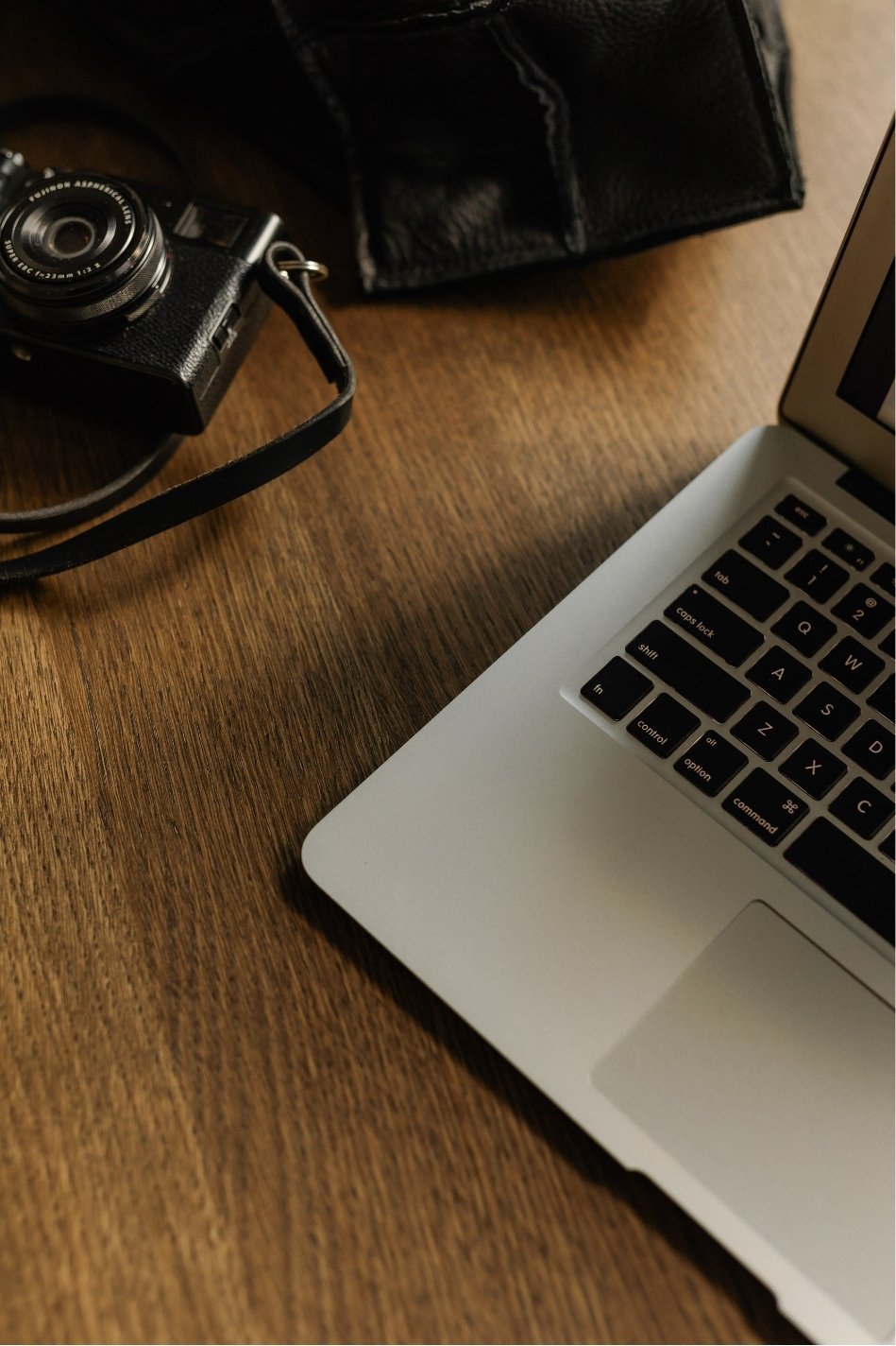 Part of a laptop on a wooden desk, with a camera and a black leather bag nearby.