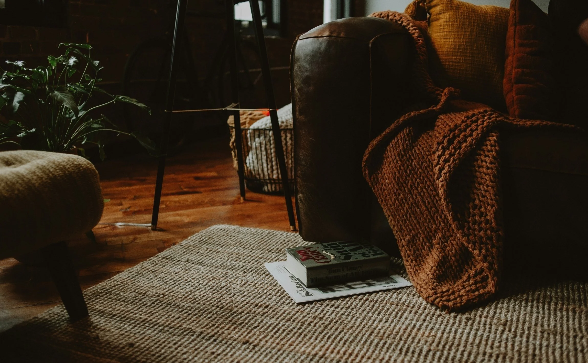 Cozy living room corner with a dark leather sofa draped with a chunky knit blanket, a patterned rug on wooden floor, a small stack of books and magazines on the floor near the sofa, a large plant on the left, and a woven basket in the background.