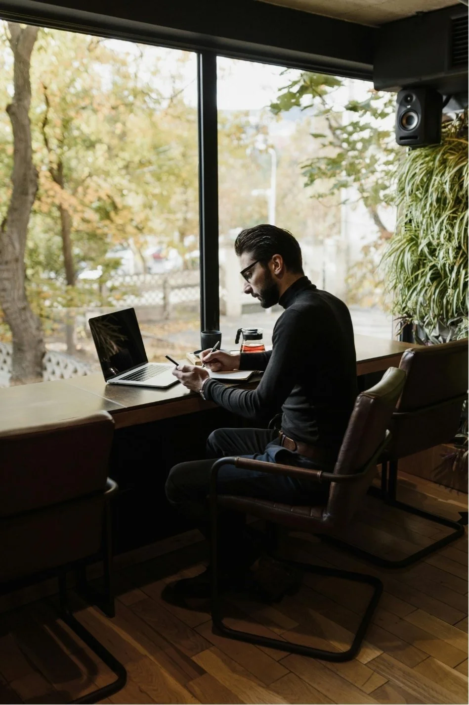 Man sitting at a wooden table near a large window, looking at his phone, with a laptop and a glass teapot on the table, in a cozy indoor setting with plants and trees outside.