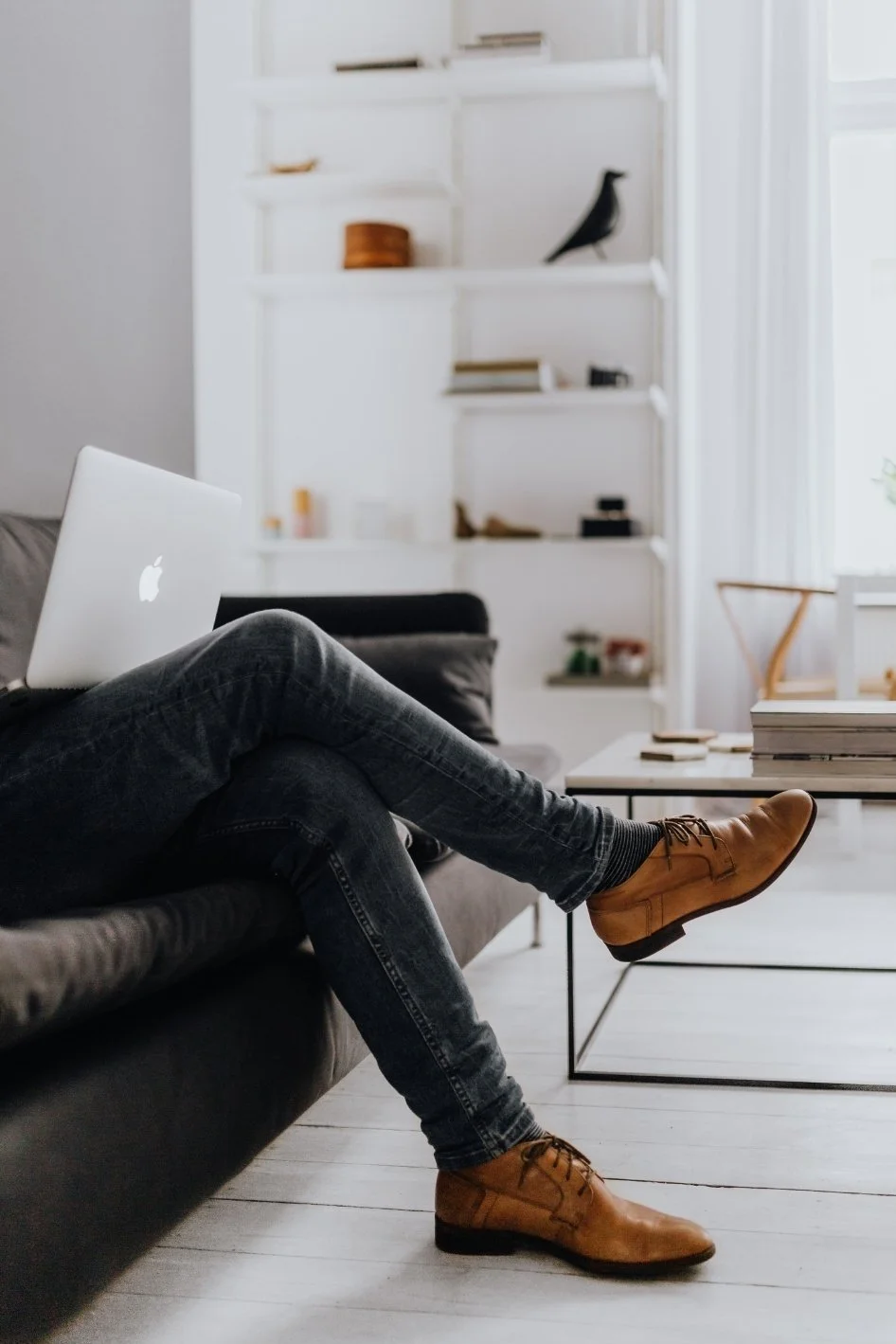 Person wearing brown shoes and dark jeans sitting on a black sofa with crossed legs, using a silver laptop, in a minimalistic, well-lit living room with white walls and shelves.