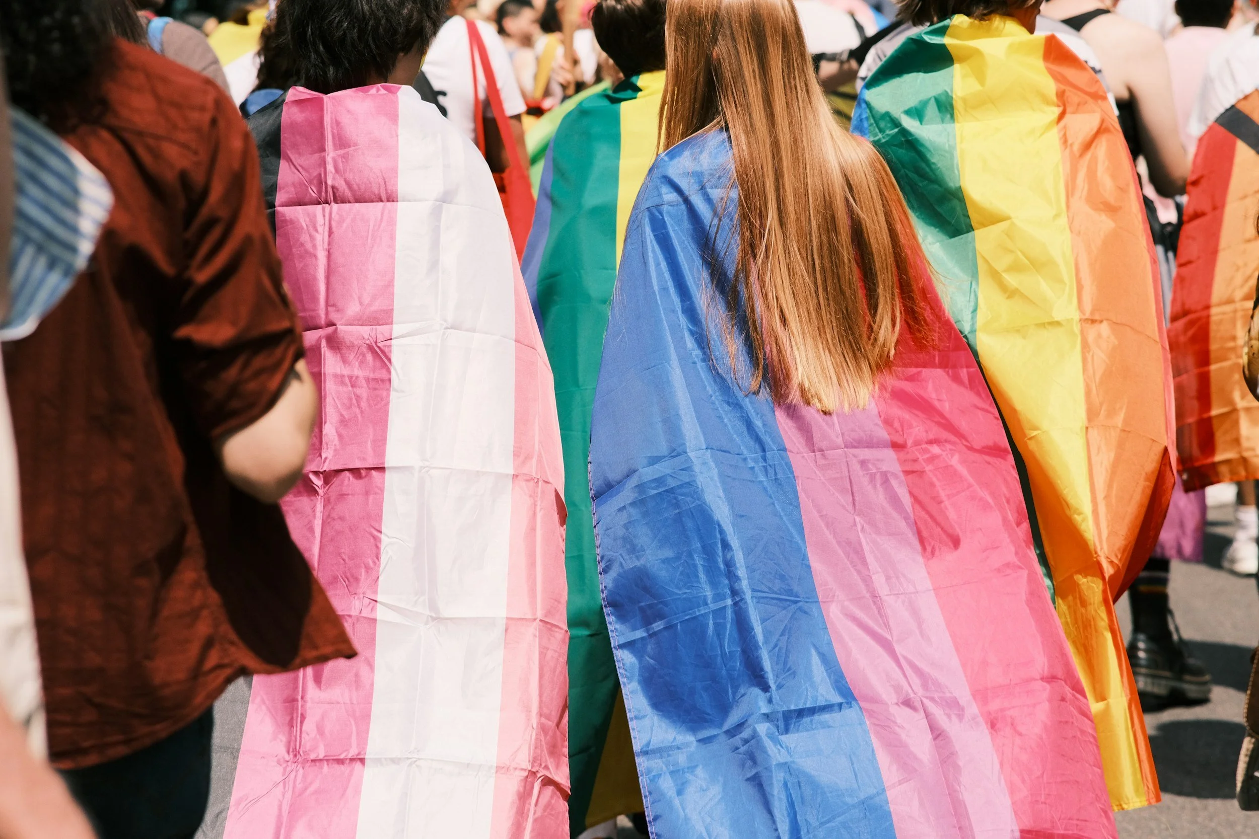 People at a pride parade wearing rainbow-colored flags as capes.