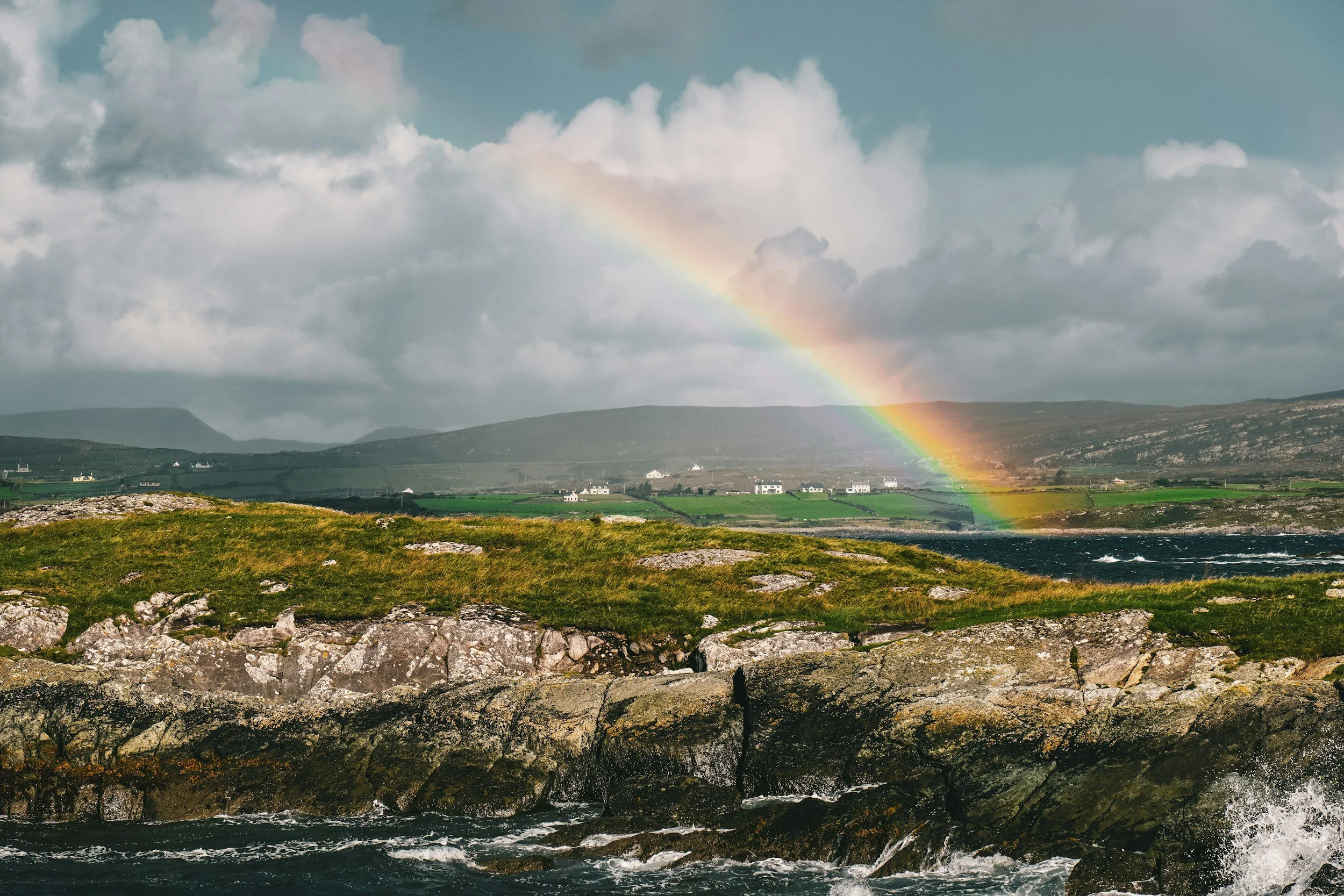 A landscape featuring a rainbow arching over a grassy shoreline with rocks and the ocean, under a partly cloudy sky.
