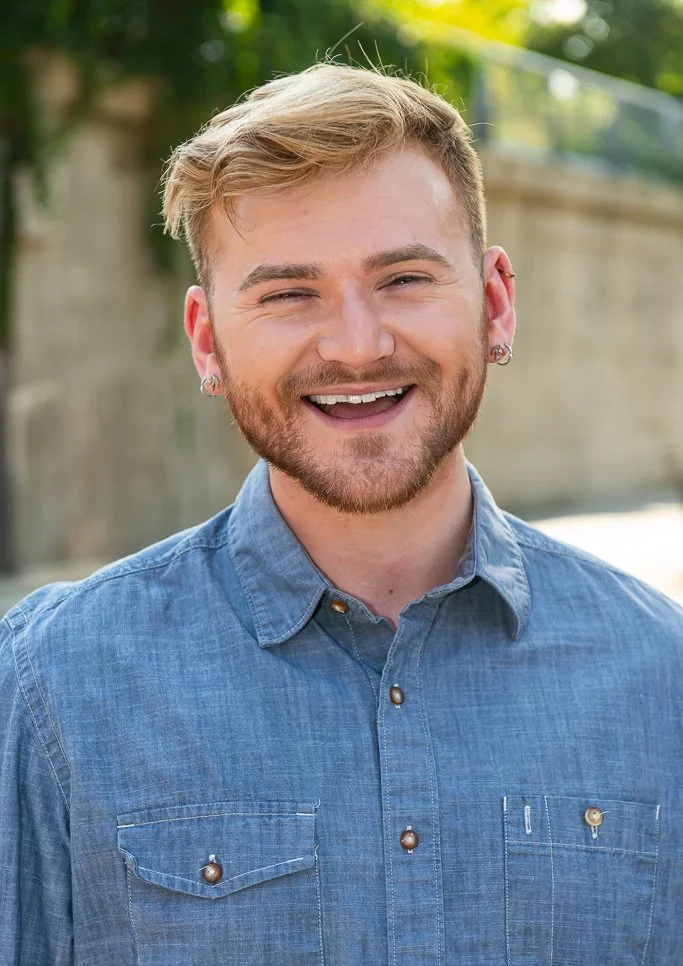 A young man with blonde hair, a beard, and earrings, smiling outdoors in front of a wooden fence and greenery, wearing a blue denim shirt.