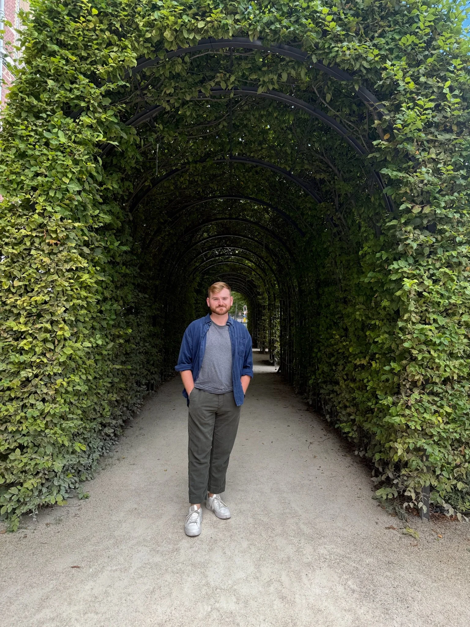 A man standing in a tunnel of green foliage with his hands in his pockets, wearing a gray shirt, dark green pants, a blue jacket, and white sneakers.