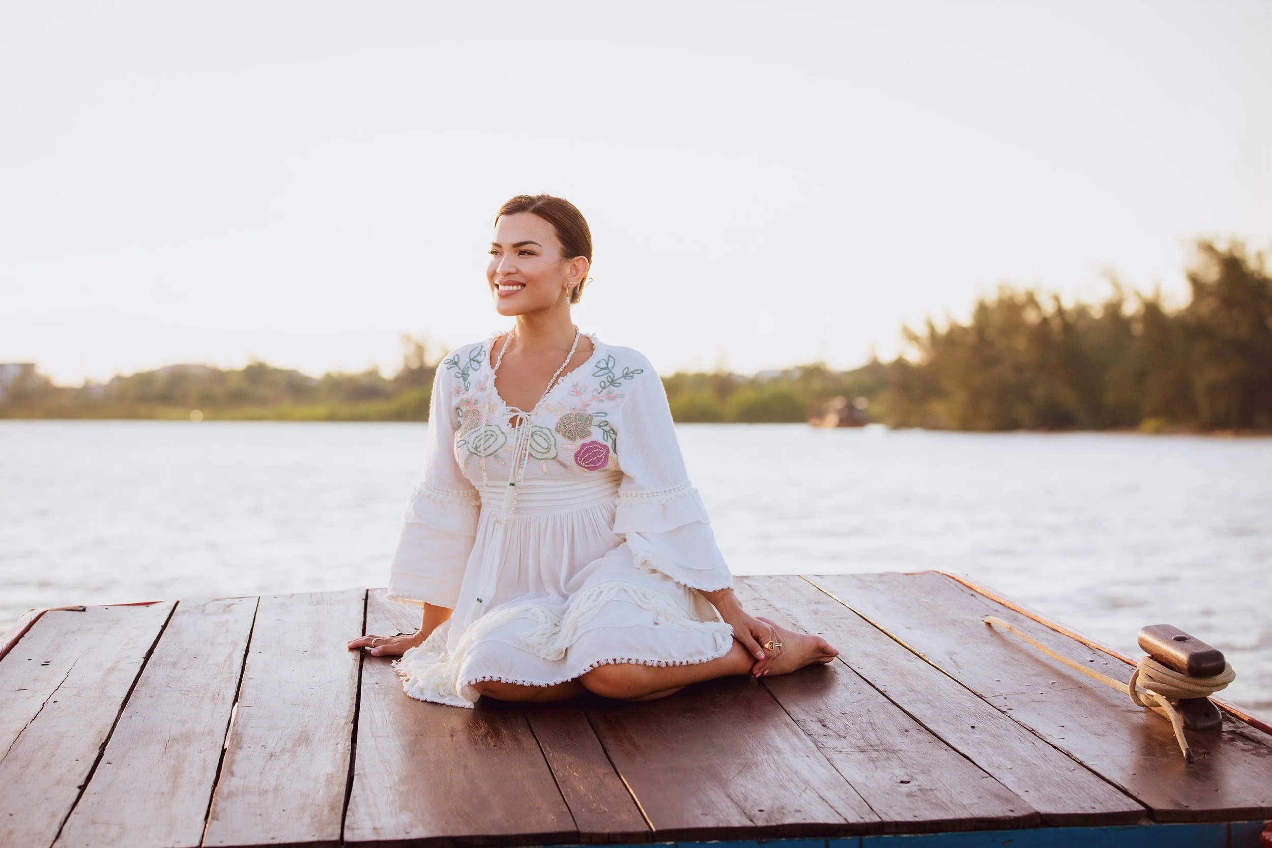 A woman in a white embroidered dress is kneeling on a wooden dock by a body of water, smiling and looking to her right during sunset or sunrise.