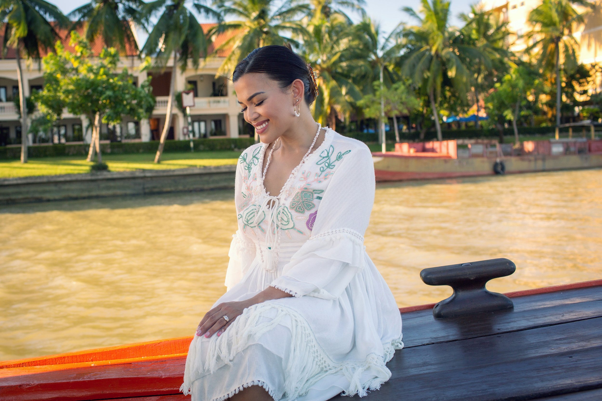 A woman in a white embroidered dress sitting on a boat by the water, smiling with her eyes closed, with palm trees and buildings in the background during the day.
