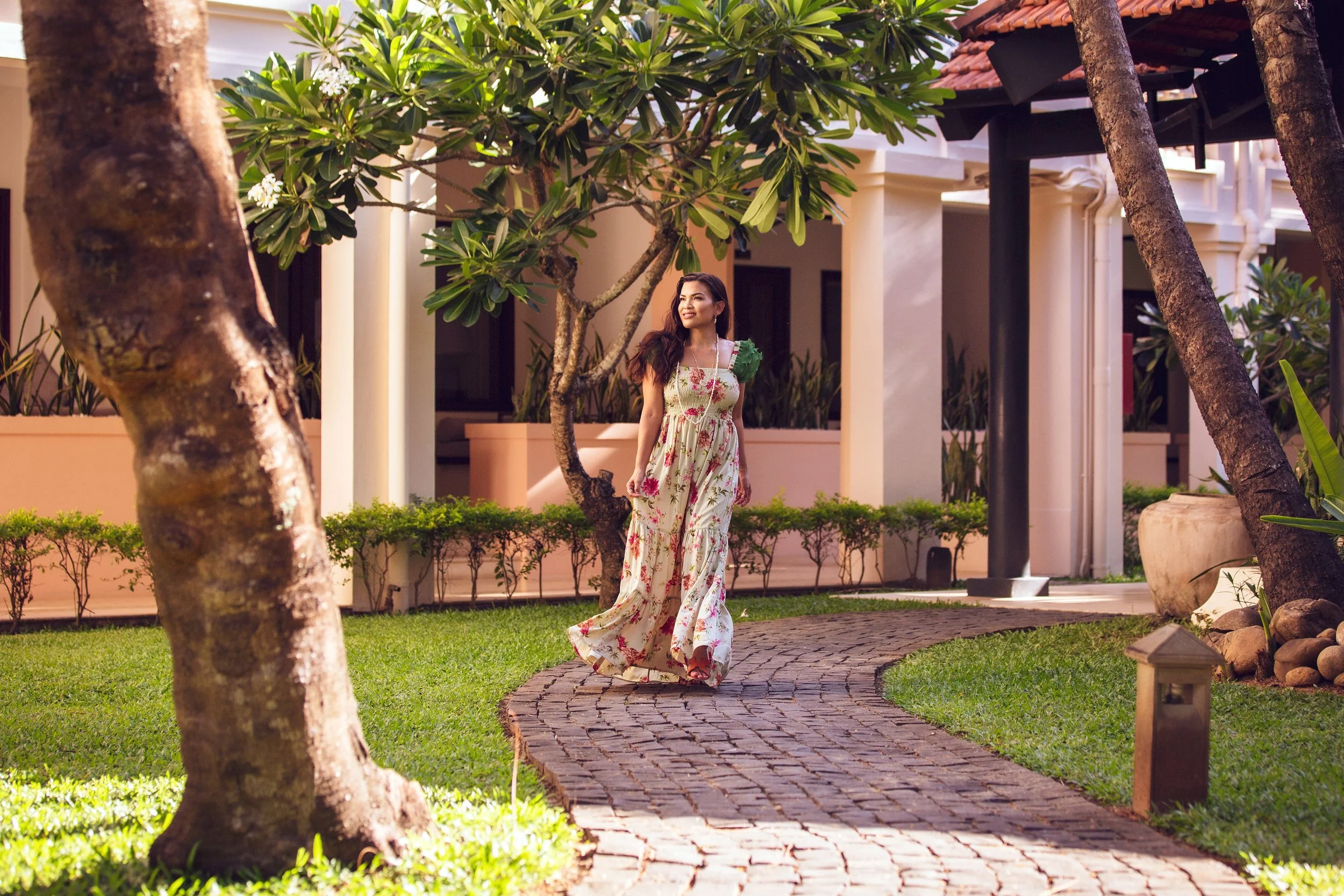 A woman in a floral dress walking along a curved brick pathway in a lush garden area outside a house with a porch and large windows.