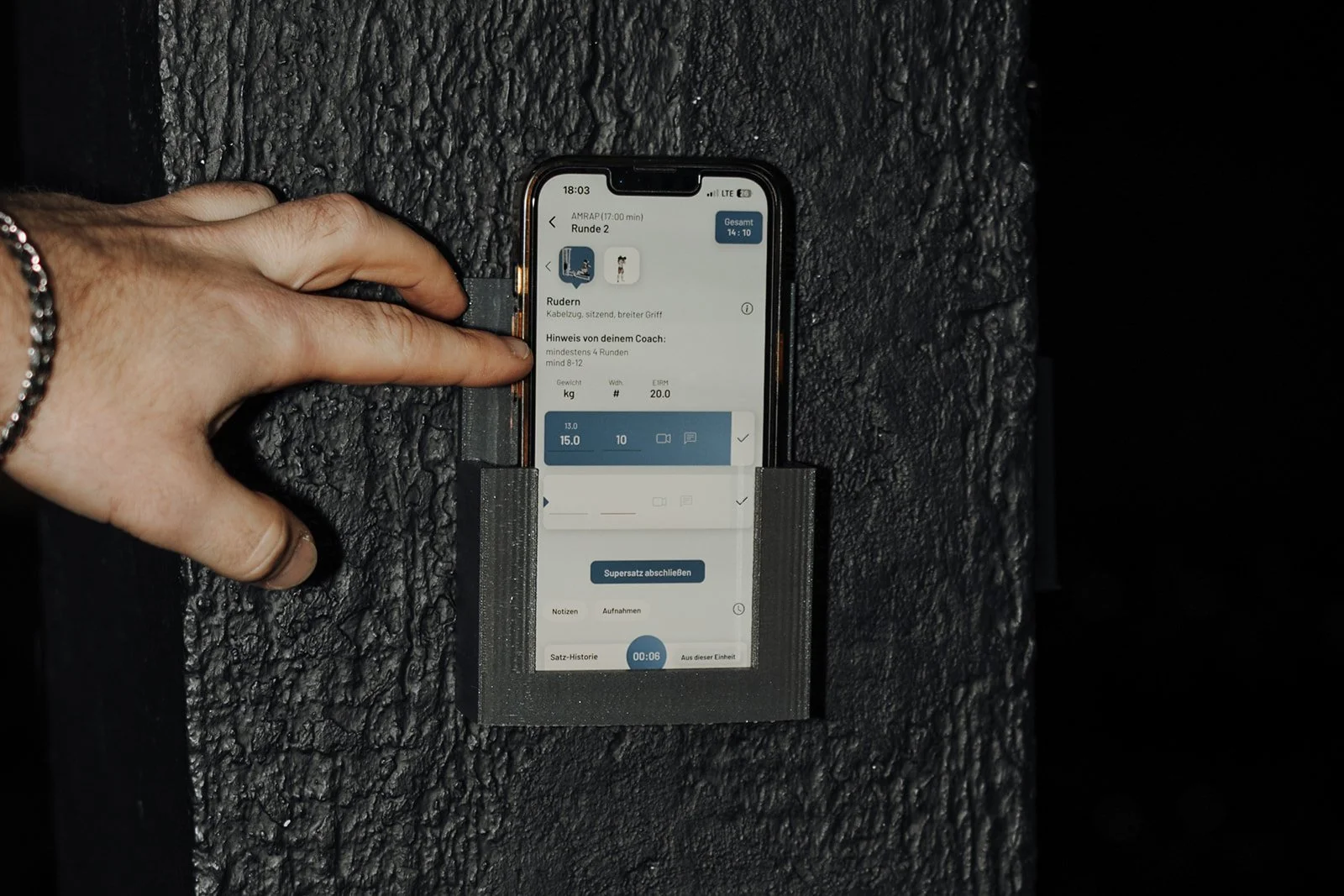 Hand hält Smartphone in einer schwarzen Halterung an einer schwarzen rauen Oberfläche, das Display zeigt eine Fitness-App mit Trainingseinheit.