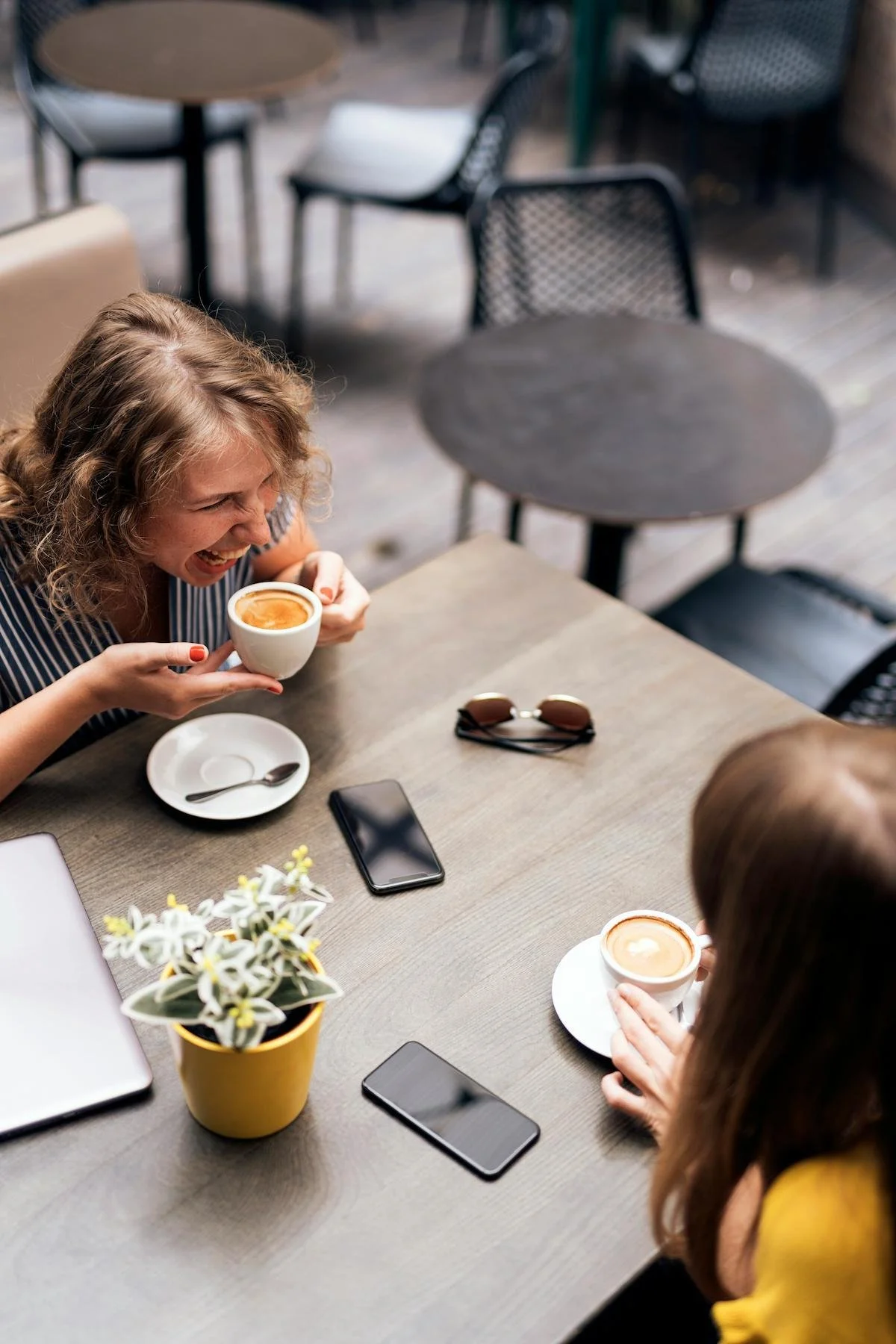 Two women sitting down for coffee together. One woman with curly blond hair is laughing and leaning forward as they talk over the table.