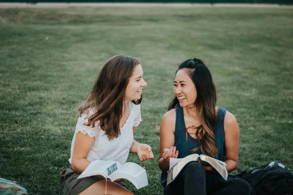Two female friends laughing in a grassy area while sitting on a blanket together.