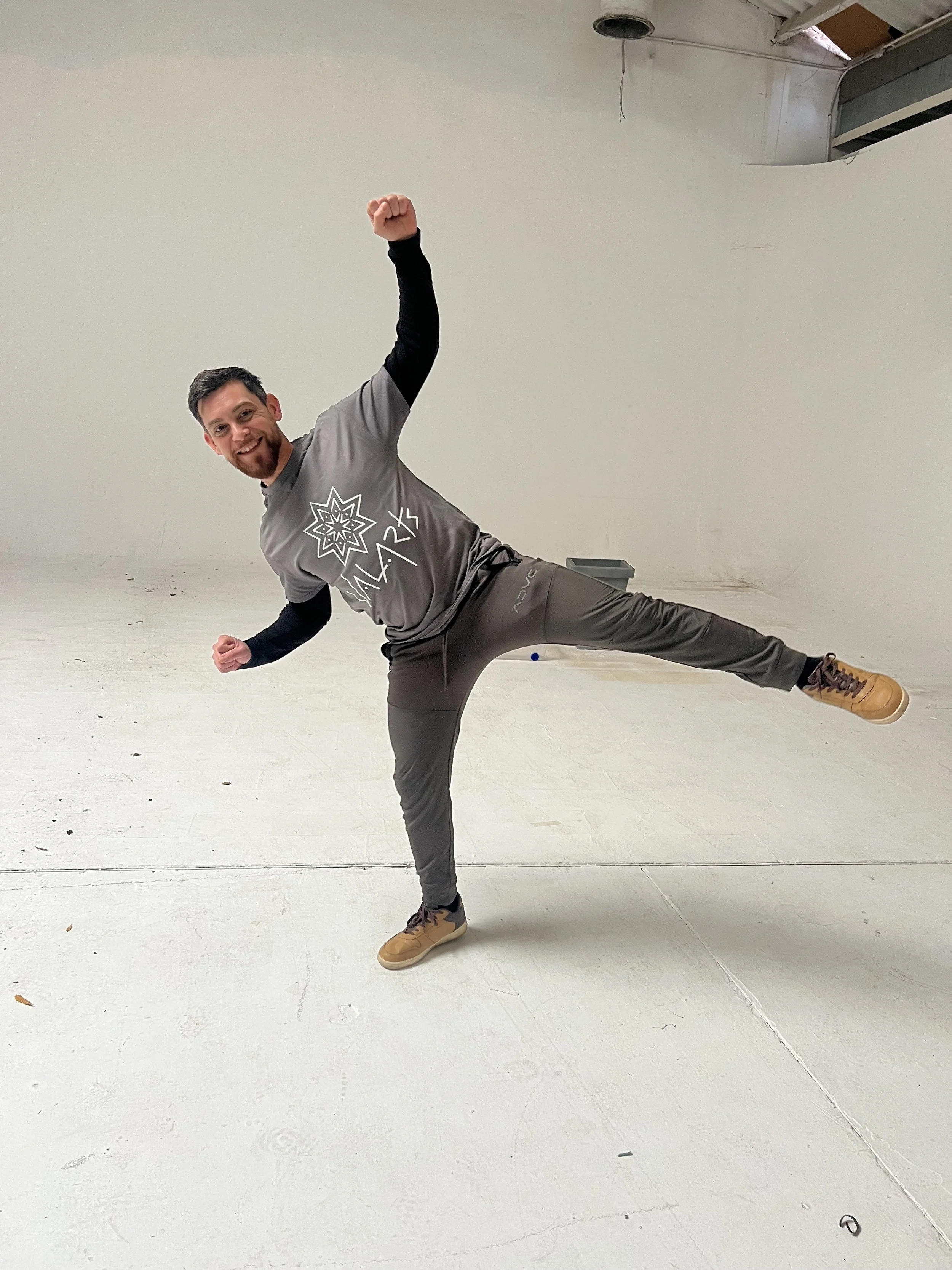 A man wearing a ValArts shirt and smiling, balancing on one leg with the other leg extended sideways, in a dance pose inside an empty white studio.