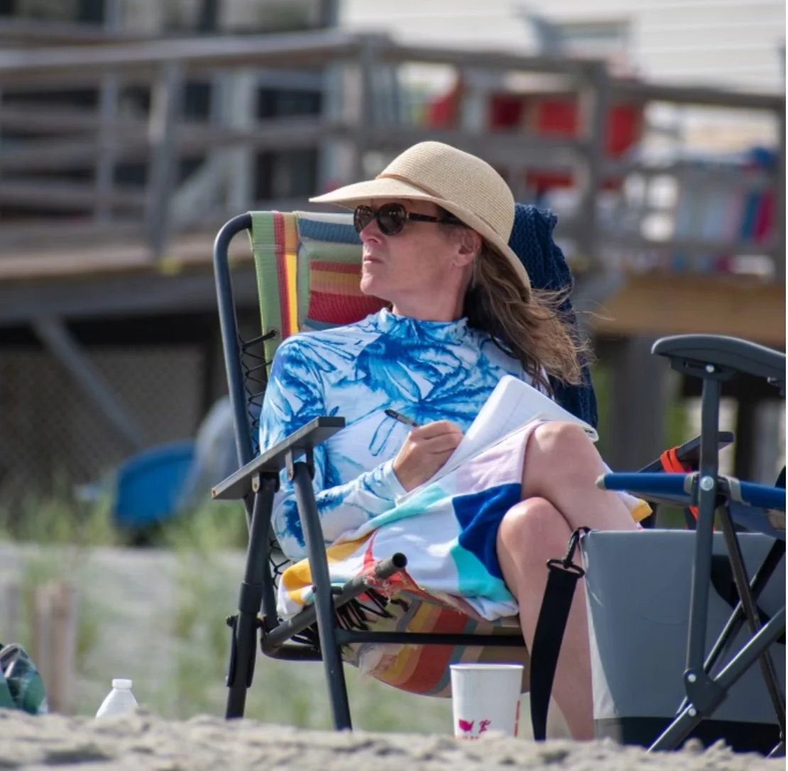 Woman sitting on beach chair, wearing a large sun hat and sunglasses, writing in a notebook, with a towel draped over her legs, near a beach with sand and some equipment in the background.