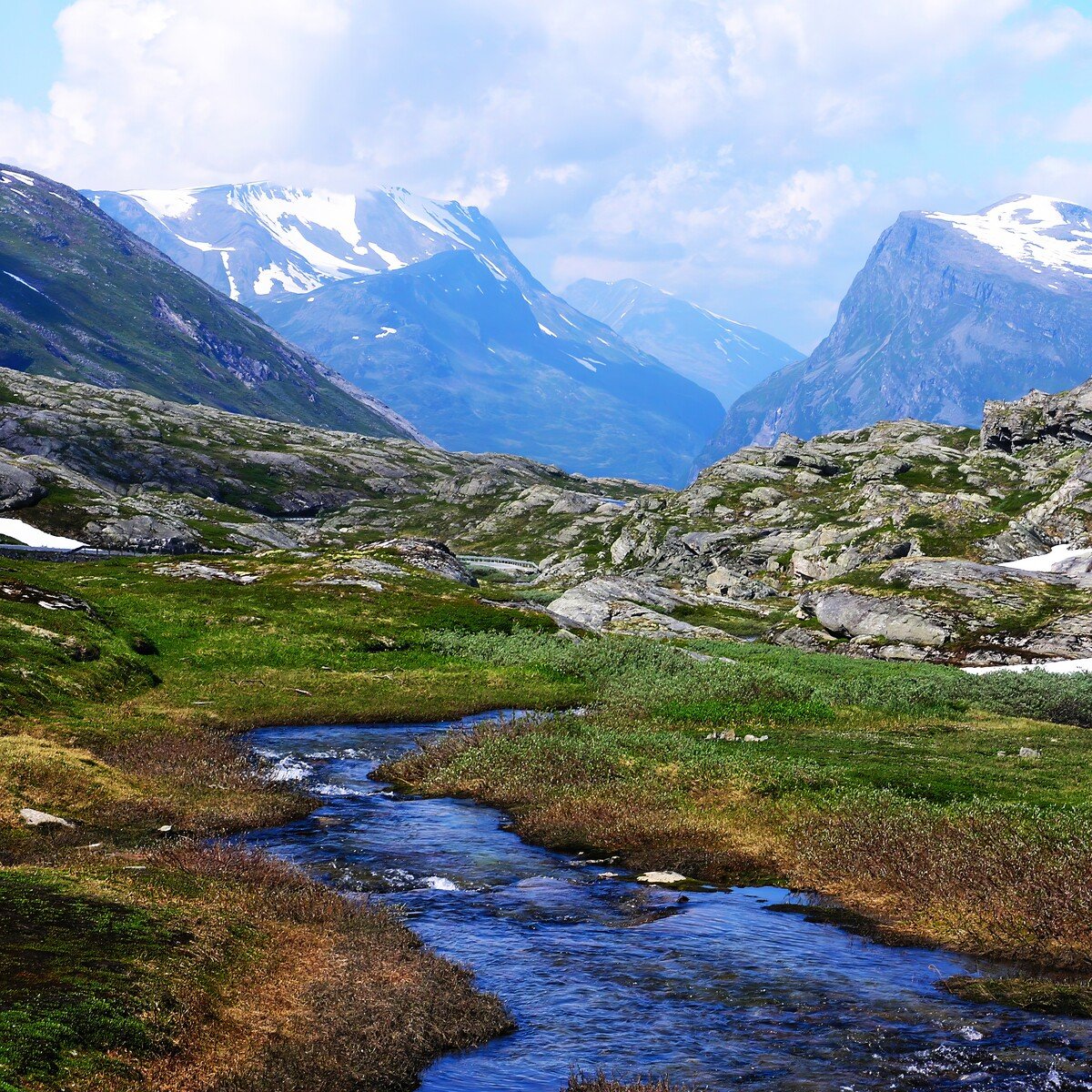 A mountain valley with a small stream flowing through green terrain, surrounded by rocky slopes and snow-capped peaks under a partly cloudy sky.