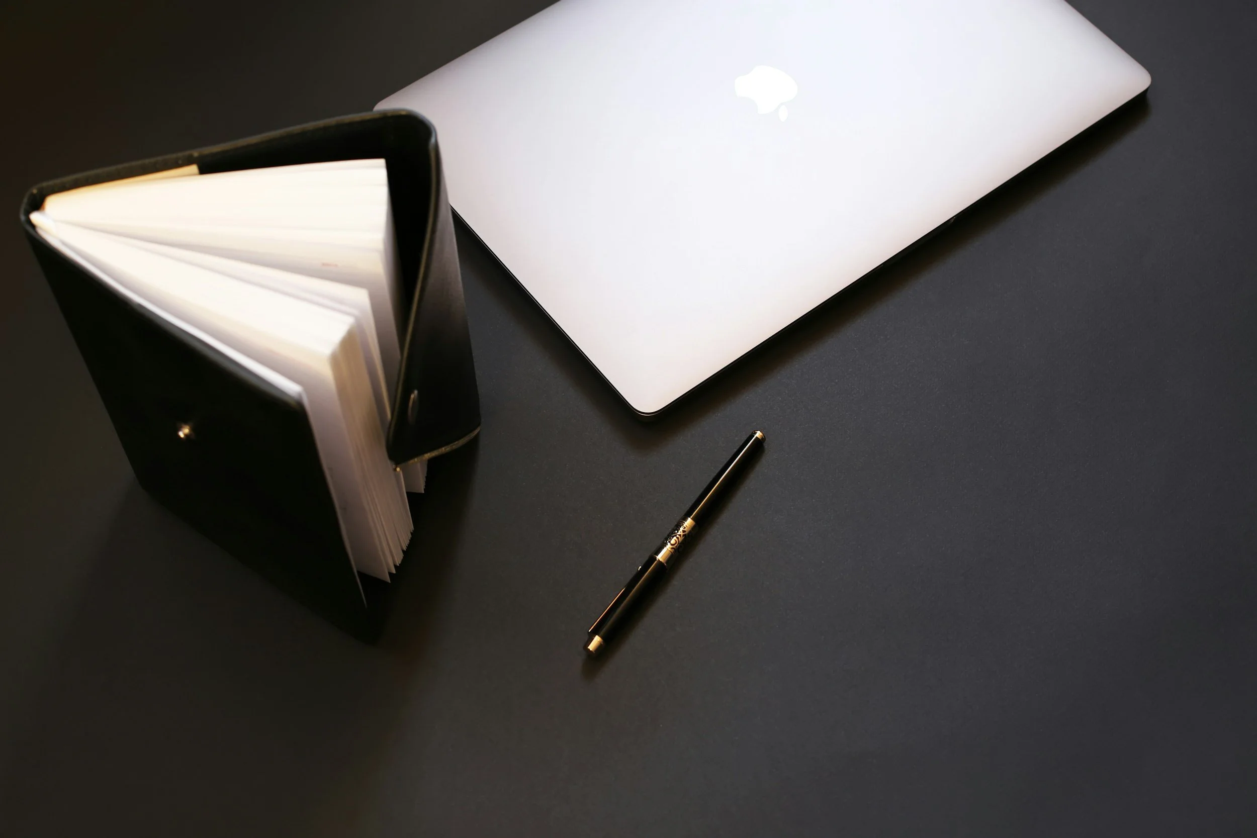 A laptop, a black notebook with a gold pen on a black surface, and an open black planner with blank pages.