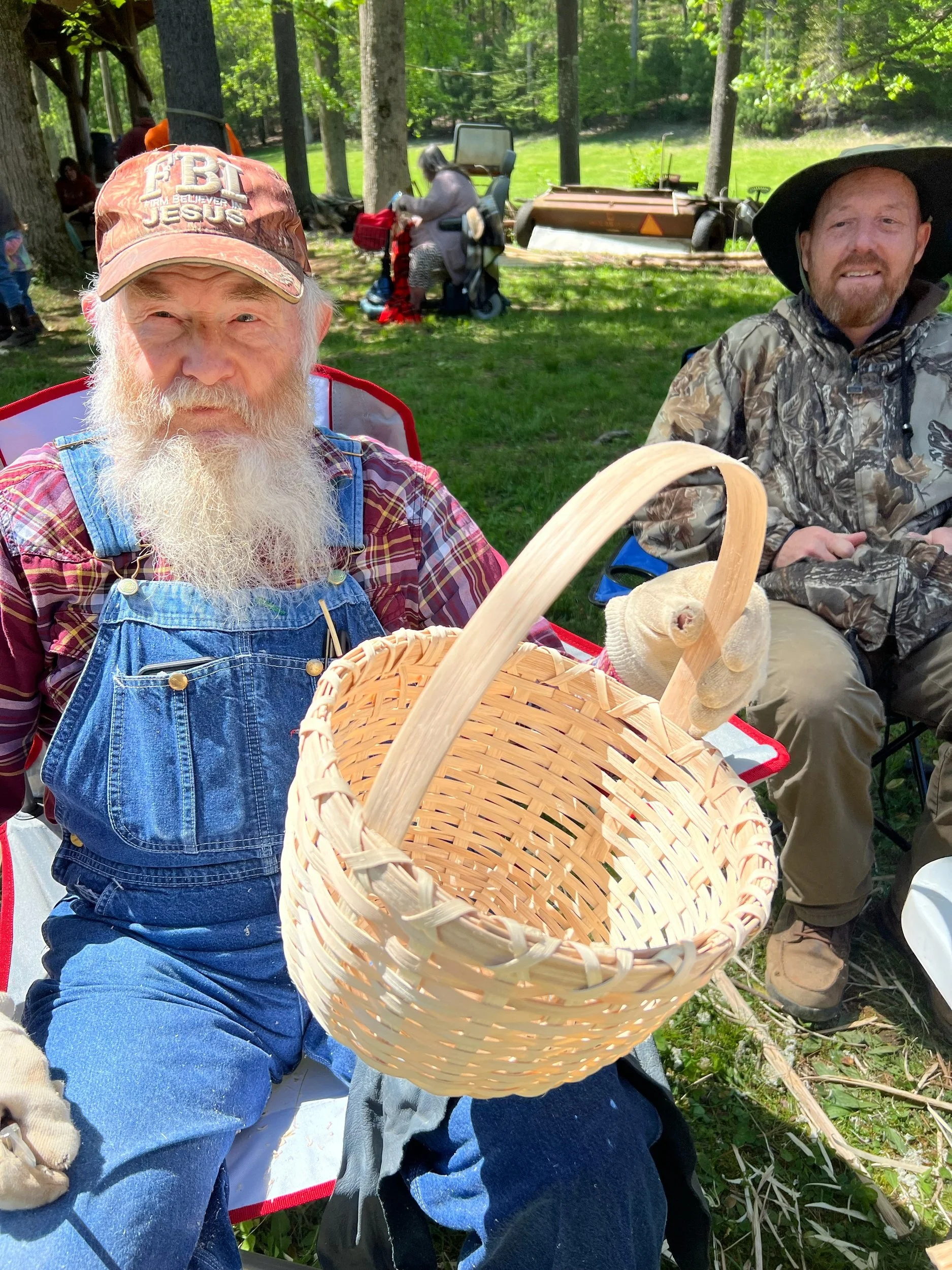 An elderly man with a long white beard wearing a red plaid shirt, blue denim overalls, and a cap that says "FBI Believers in Jesus," holding a wicker basket and sitting outdoors in a wooded area. Another man with a beard wearing camouflage jacket and