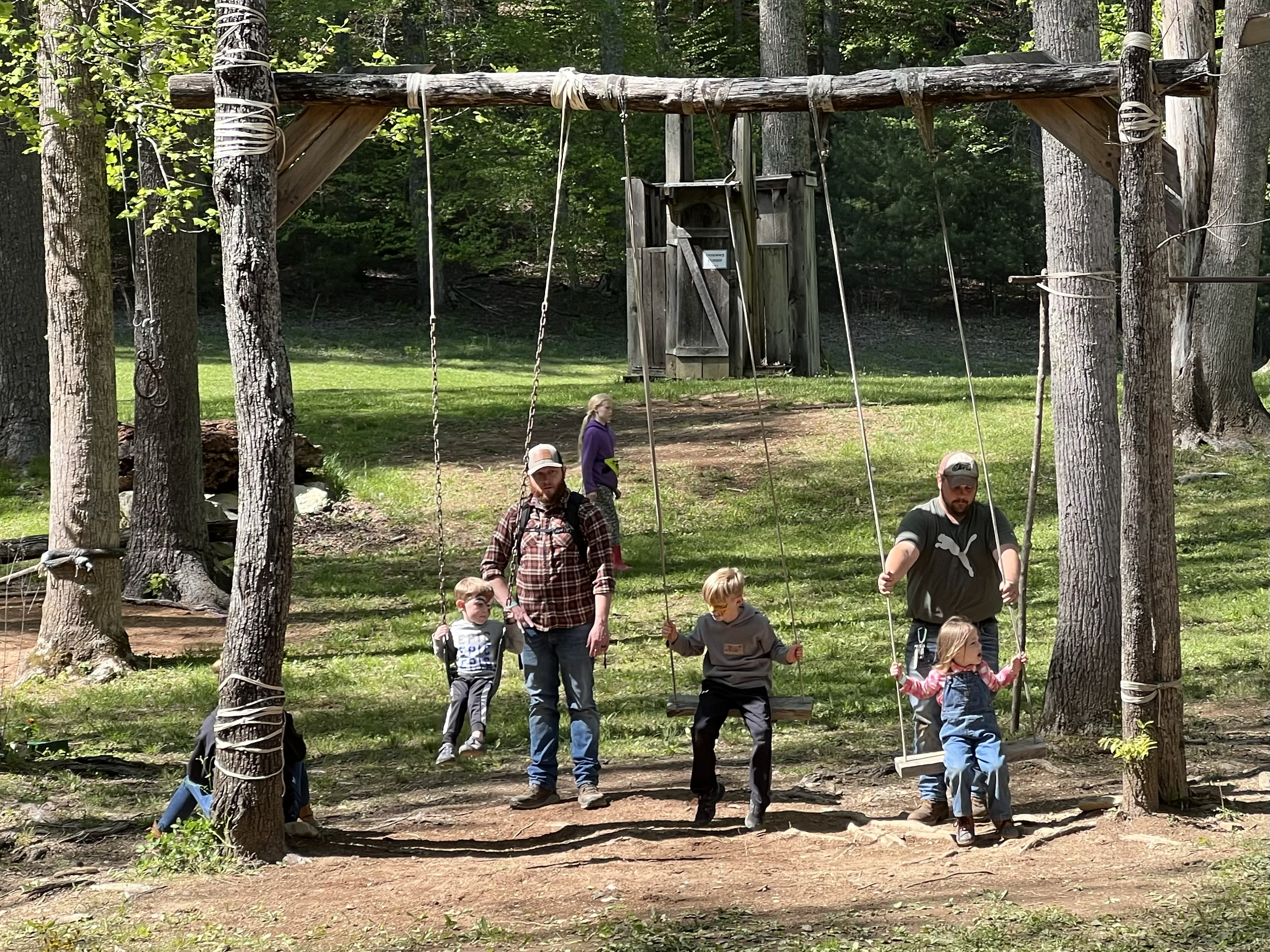 Two adults and four children at a playground in a wooded area, with two swings hanging from a wooden frame, some trees, and a small wooden playhouse in the background.