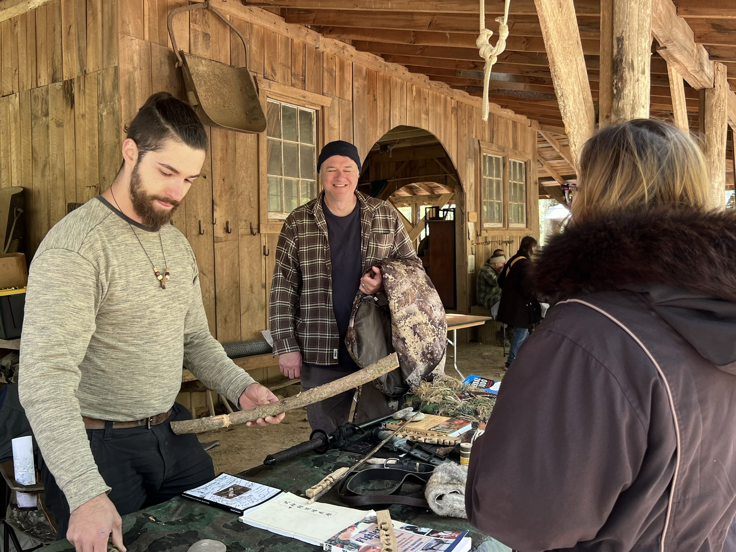 A man with a beard and long-sleeve shirt holding a wooden stick at a table, smiling man in plaid shirt and black beanie behind him, and a woman with glasses and dark jacket facing them in an indoor rustic wooden setting.