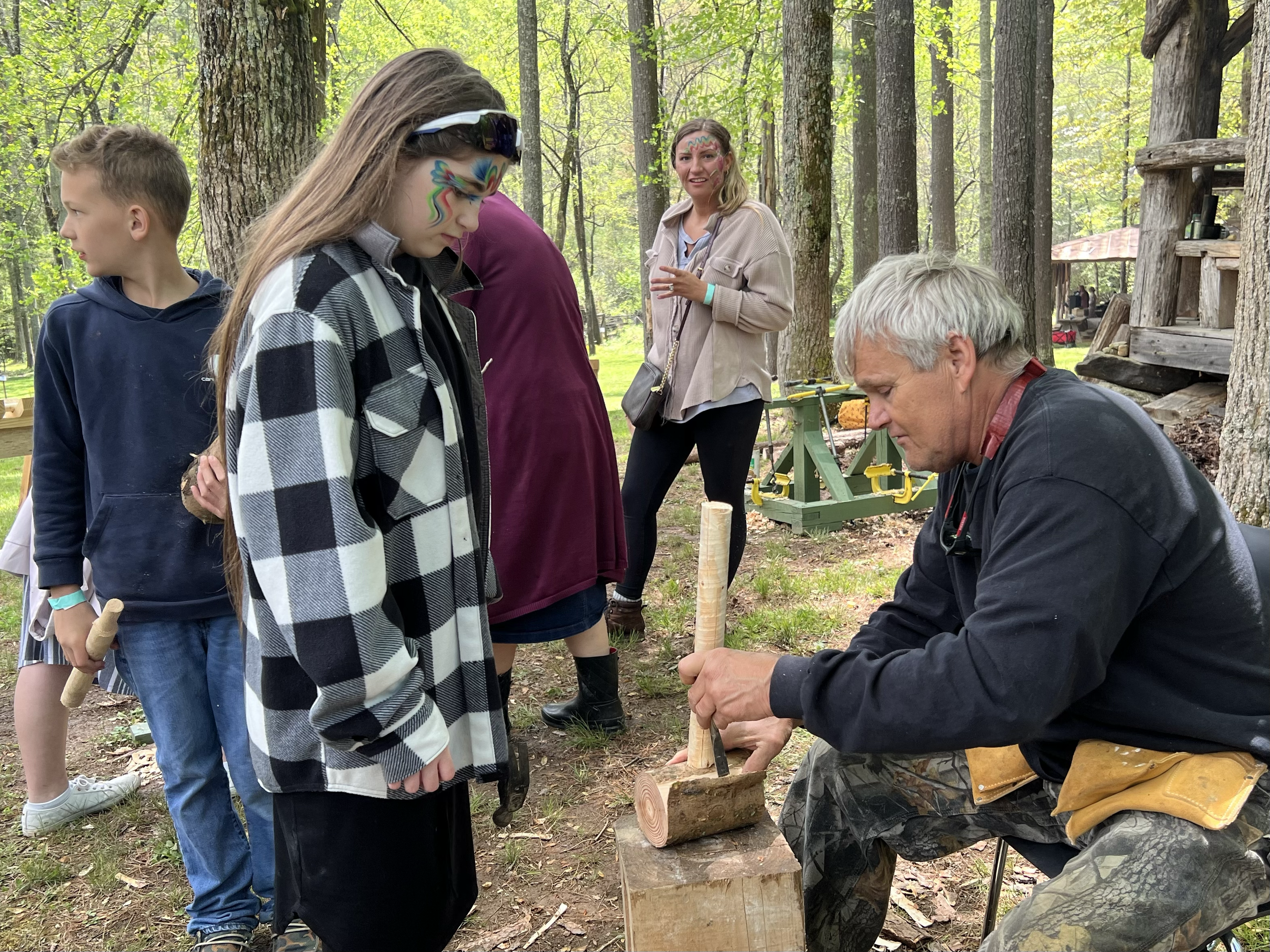 An elderly man demonstrating wood carving to a group of children and women in a forest during daytime. The man is holding a chisel and working on a wooden log, while the children and women watch attentively.